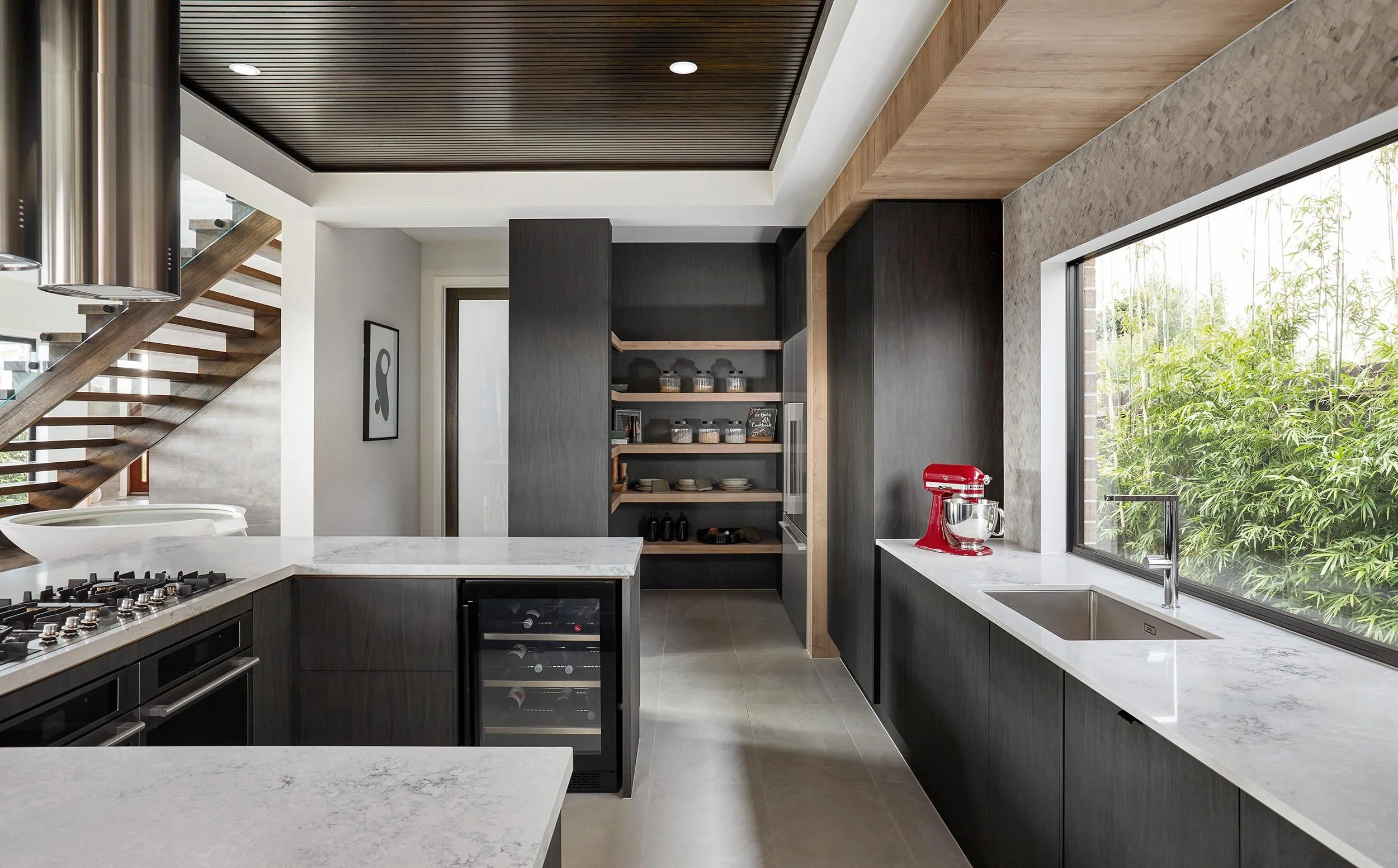 Modern kitchen with dark cabinetry, marble countertops, a large window overlooking green foliage, and a red stand mixer on the counter.