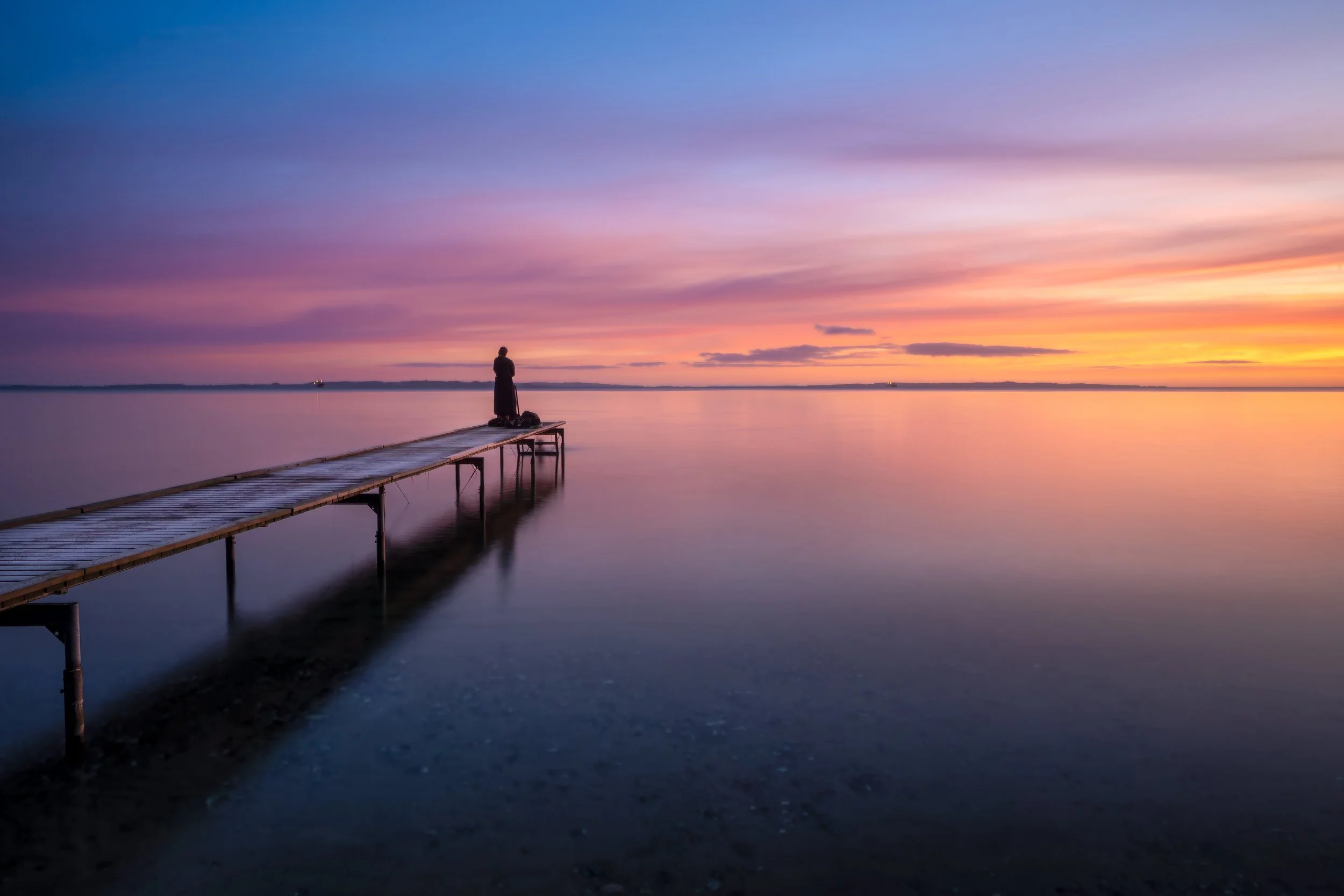 A person standing on a dock by a calm body of water during sunset with a colorful sky.