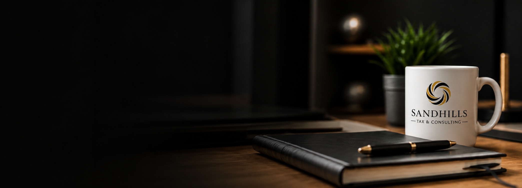 Desk with Sandhills Tax & Consulting mug, black notebook, gold and black pen, potted plant, and bookshelf in dim lighting.