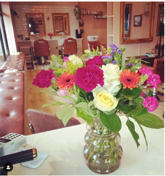 Vase with colorful flowers including pink carnations, orange gerbera daisies, white roses, and purple flowers on a table in a cozy room.