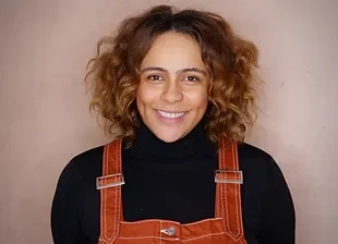 Portrait of a young woman with curly hair wearing a black turtleneck and brown overalls, smiling against a beige background.