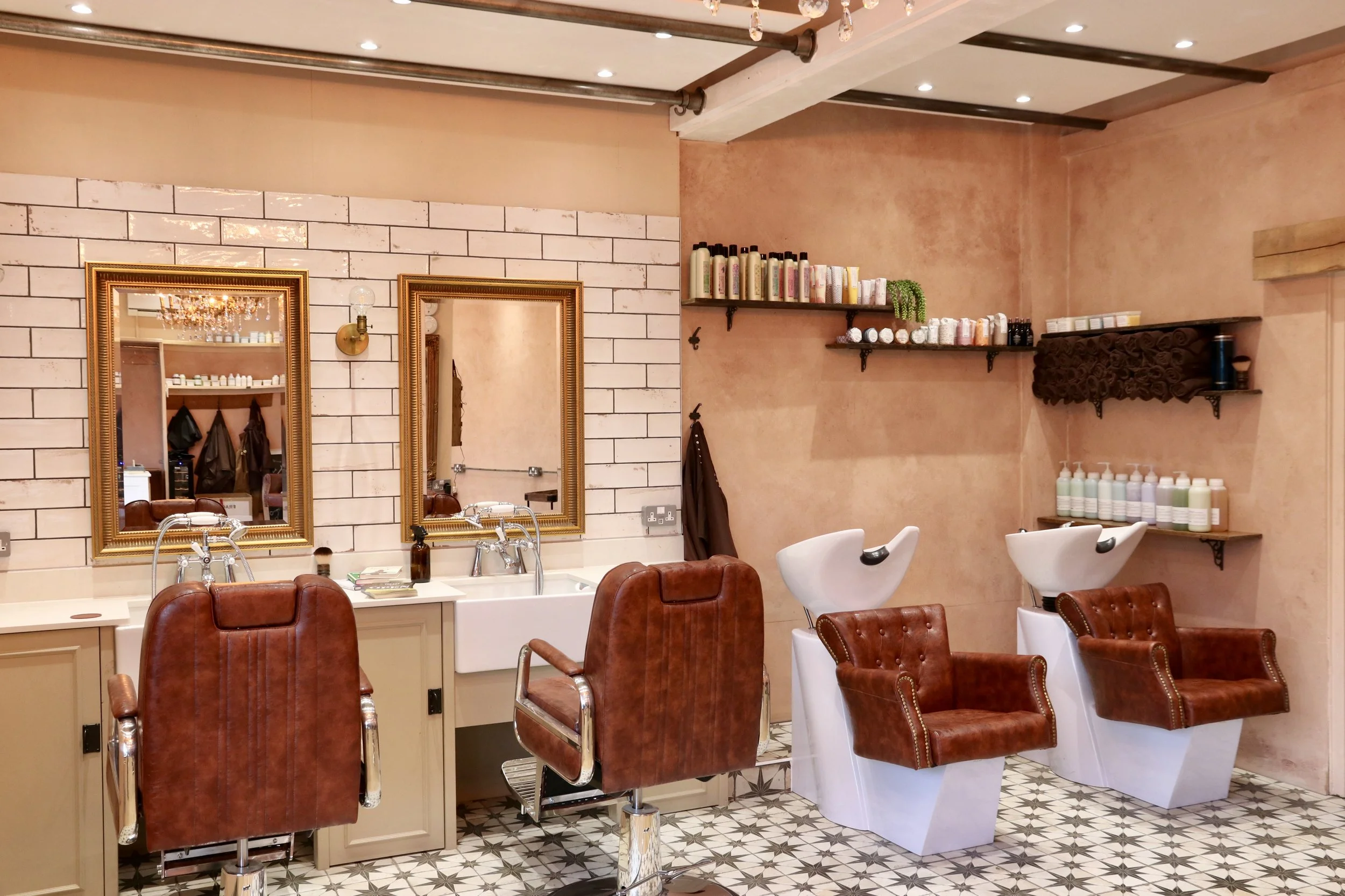 Interior of a hair salon with styling stations, salon chairs, mirrors, shelves with hair products, and white wash basins for hair washing, featuring warm beige and brown tones and decorative patterned floor tiles.