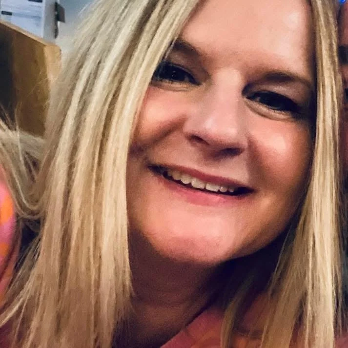 Close-up of a smiling woman with long blonde hair in an indoor setting.