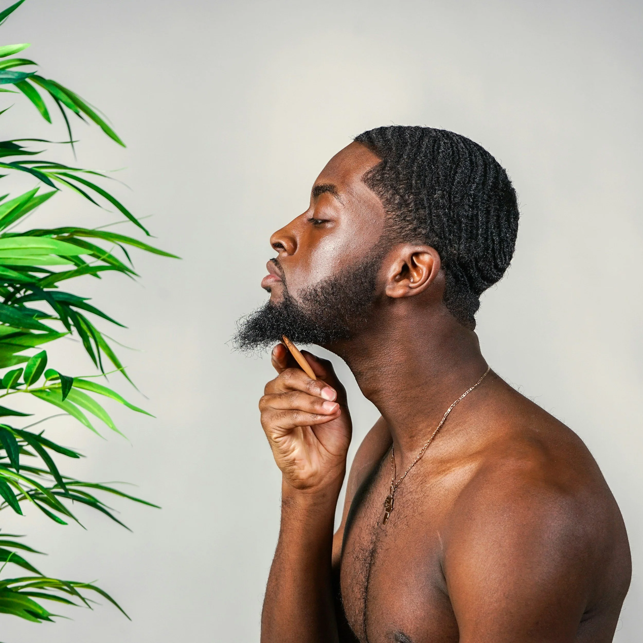 Side profile of a shirtless man with dark skin, short curly hair, and a beard, holding a pencil near his chin, with green leaves partially visible on the left and a plain light-colored background.