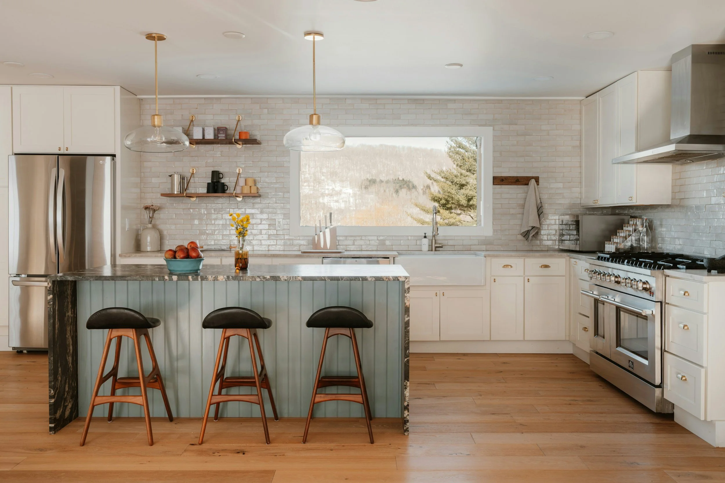 A modern kitchen with white cabinets, stainless steel appliances, a marble island with three bar stools, open shelves with cups and jars, a large window showing trees outside, and hardwood flooring.