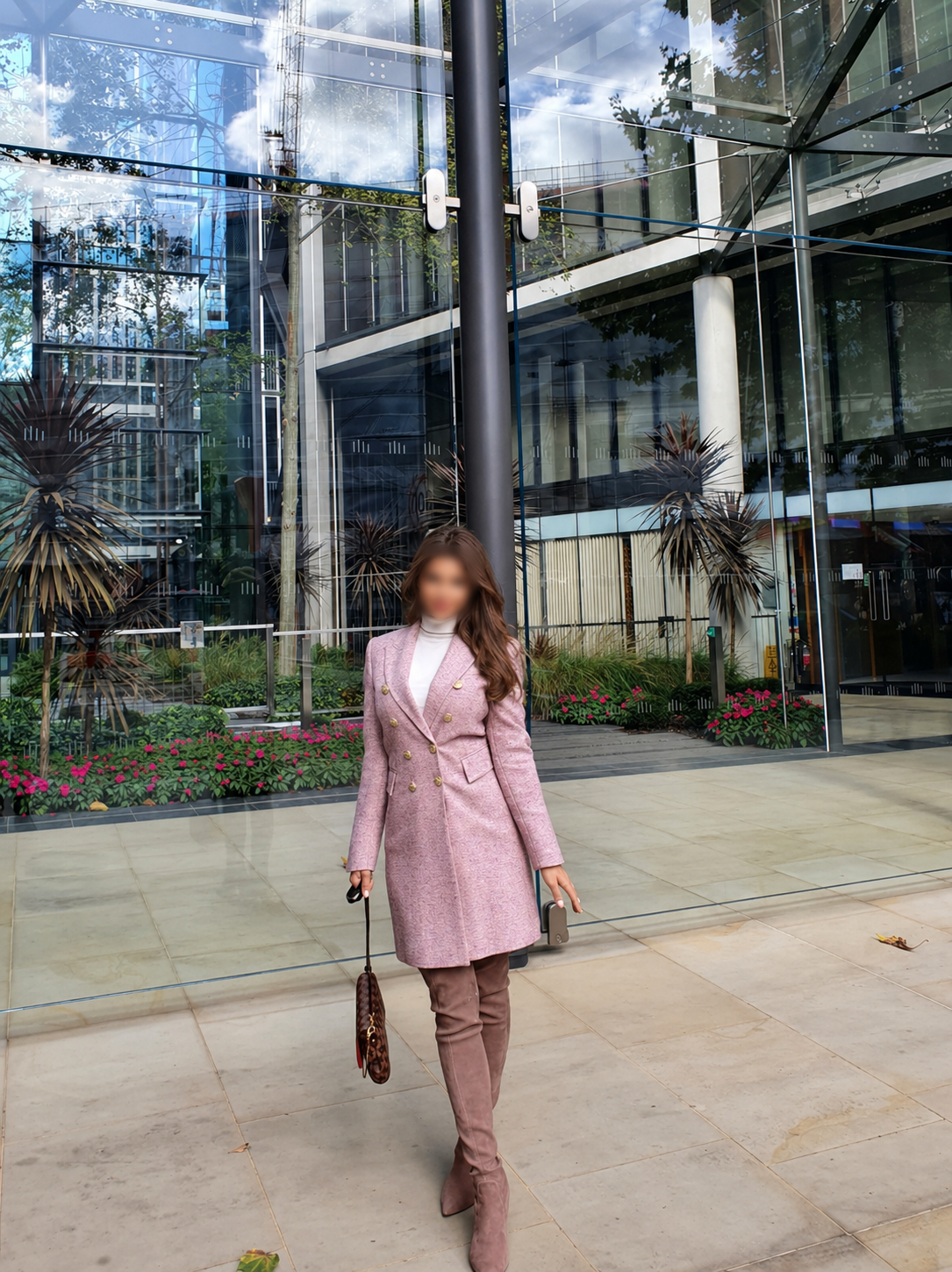 A woman in a pink coat and brown pants walking outside a modern glass building with plants and flowers in the background.