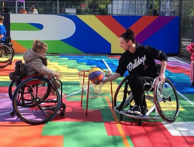 A woman in a wheelchair playing wheelchair basketball with a young girl in a wheelchair on an outdoor colorful basketball court.