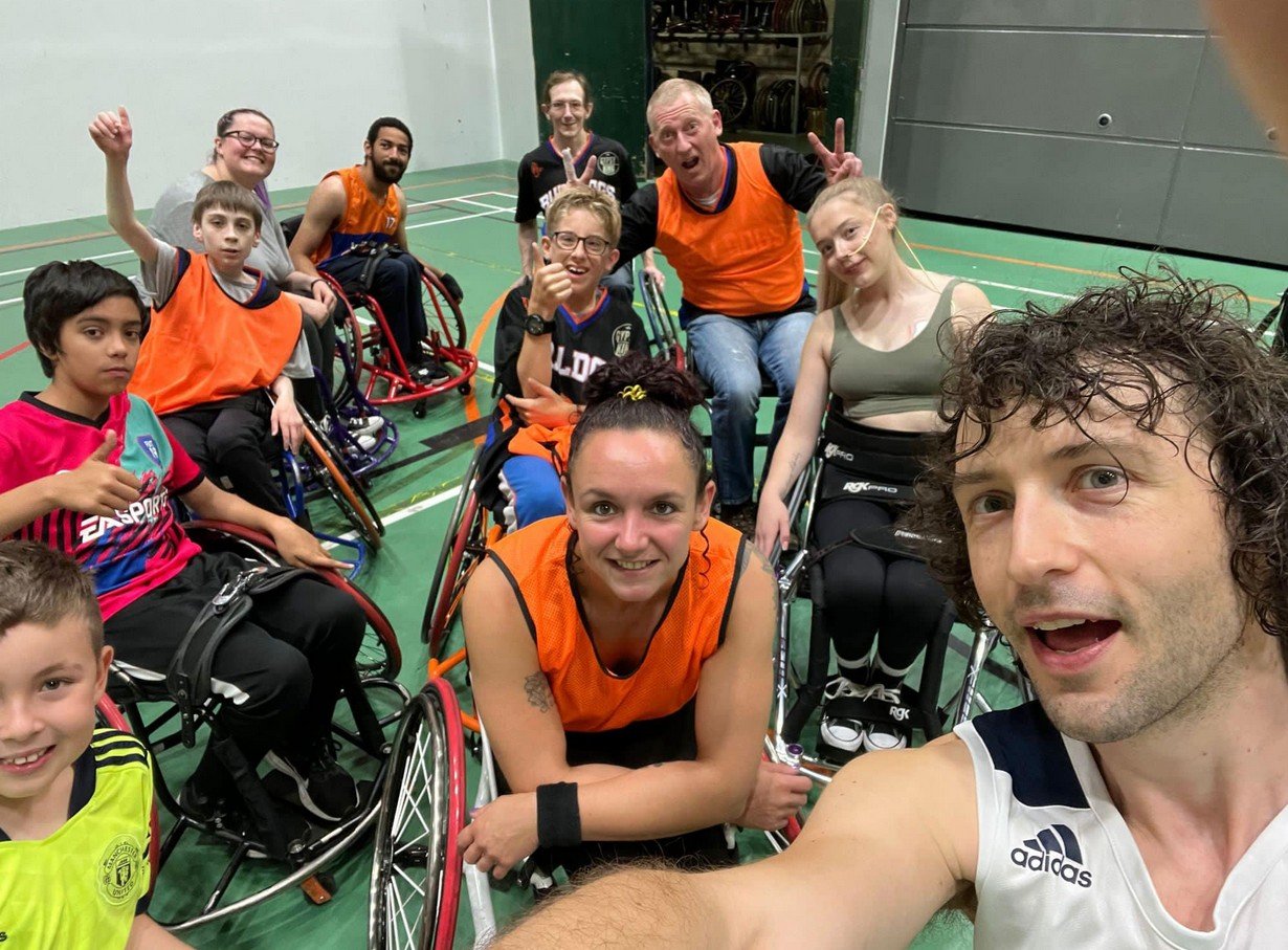 Group photo of diverse young people and adults in wheelchairs on a gymnasium court, smiling and making peace signs after a sports activity.