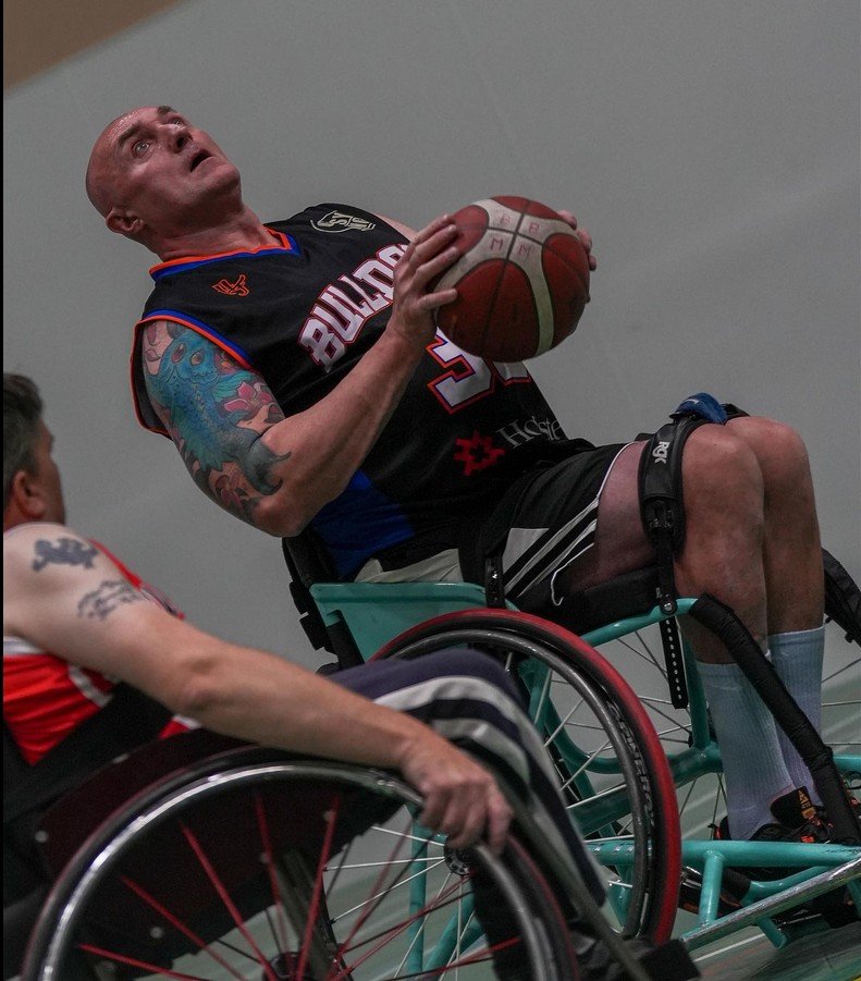 Two wheelchair basketball players compete for the ball during a game, with one player holding the ball and the other reaching for it.