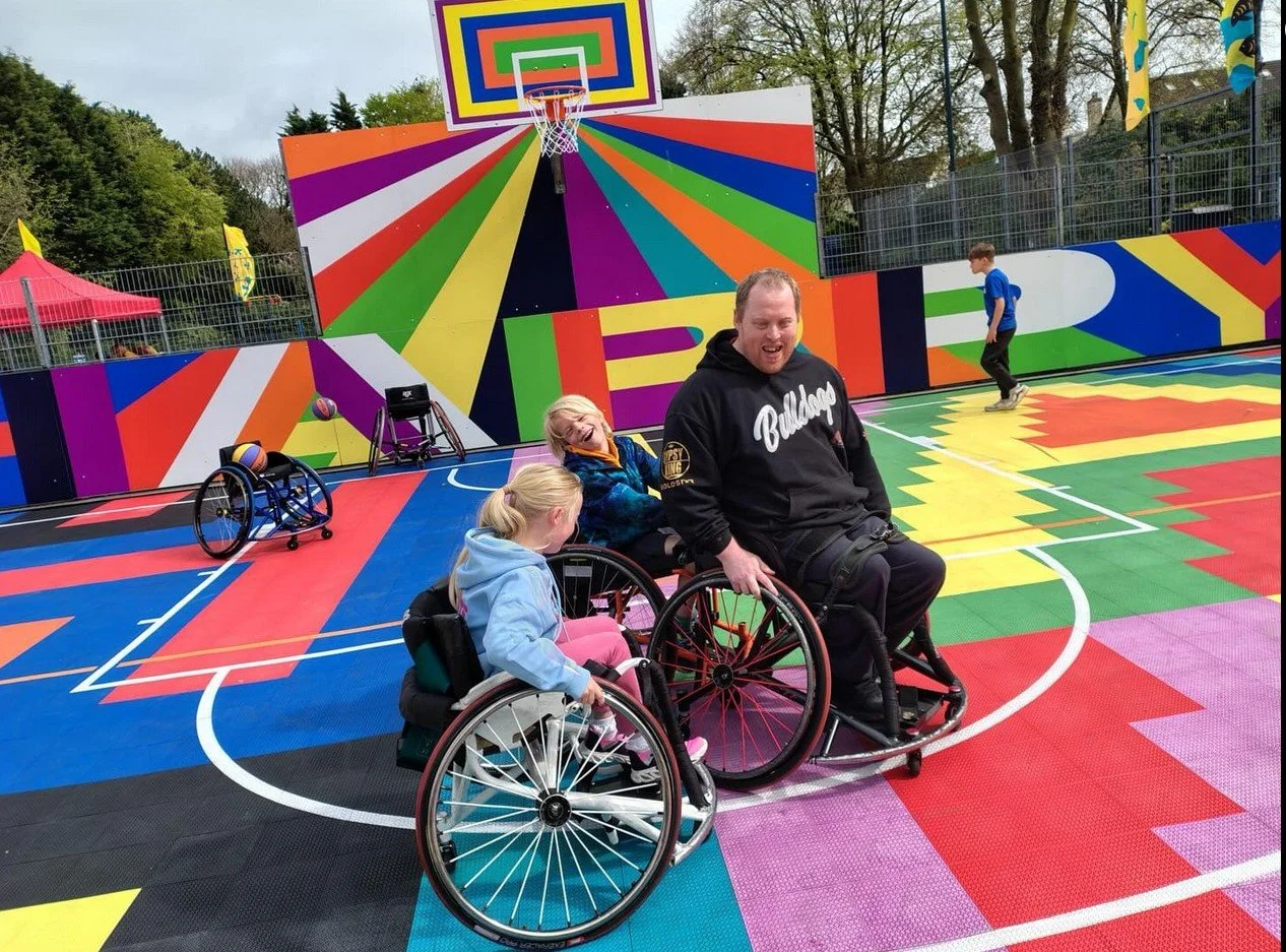 People in wheelchairs on a colorful outdoor basketball court with vibrant, rainbow-colored mural in the background.