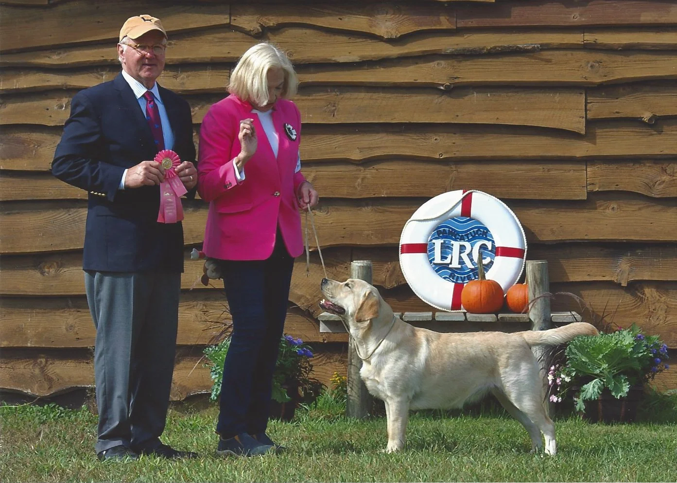 A man and a woman standing outdoors with a yellow Labrador Retriever. The man is holding a pink ribbon award, and the woman is holding a leash attached to the dog. Behind them is a wooden wall, orange pumpkins, a life preserver with the letters 'LRC,' and potted plants.