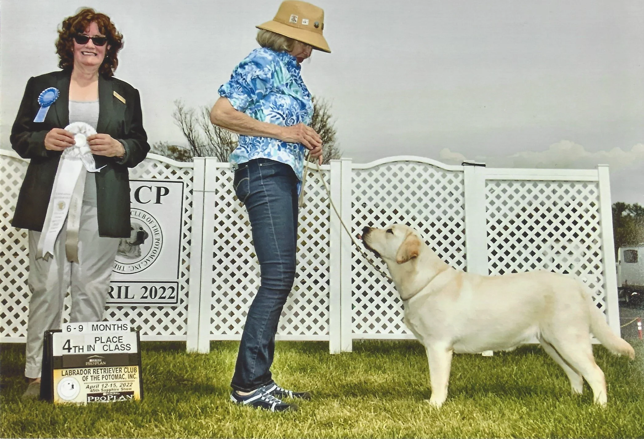 Two women standing outdoors in front of a white decorative fence, with a Labrador Retriever puppy. One woman has curly red hair, sunglasses, and a black blazer, and is holding a white ribbon. The other woman is wearing a sunhat and a blue patterned shirt, holding the puppy's leash. The puppy is sitting on the grass, looking at the woman with the hat. A sign indicates the event is for dogs aged 6-9 months, held in April 2022 at the Labrador Retriever Club of the Potomac.