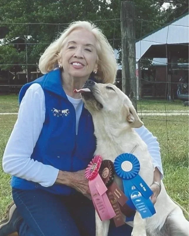 Gail Taber, wearing a blue vest and a white long-sleeved shirt, is sitting outdoors with her Labrador Retriever, which has ribbons on its chest from a show or competition. Gail is smiling, and the dog appears to be licking her face.