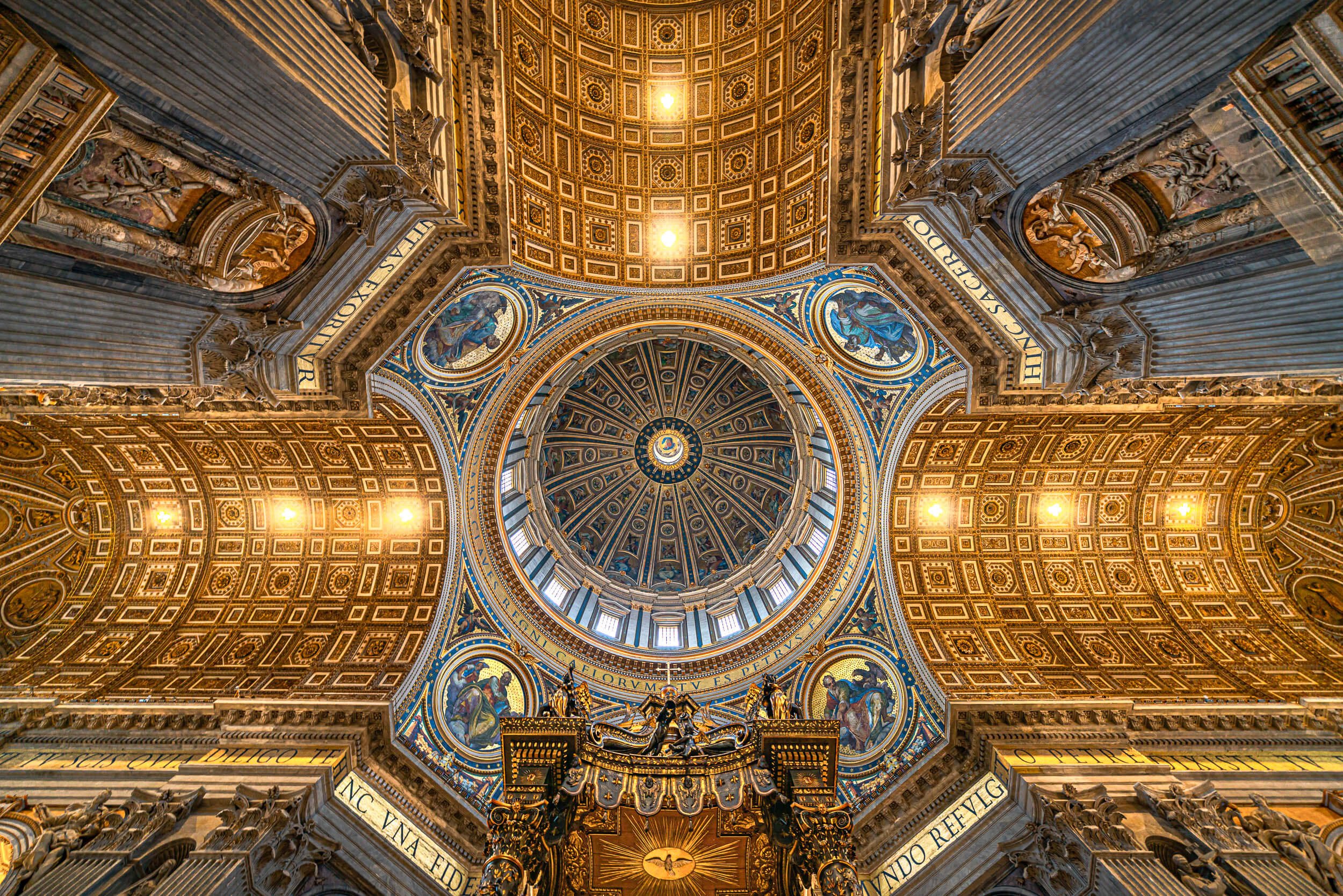 Interior view of a grand cathedral ceiling with detailed gold and blue decorative artwork, including circular and rectangular patterns, Latin inscriptions, and religious figures.