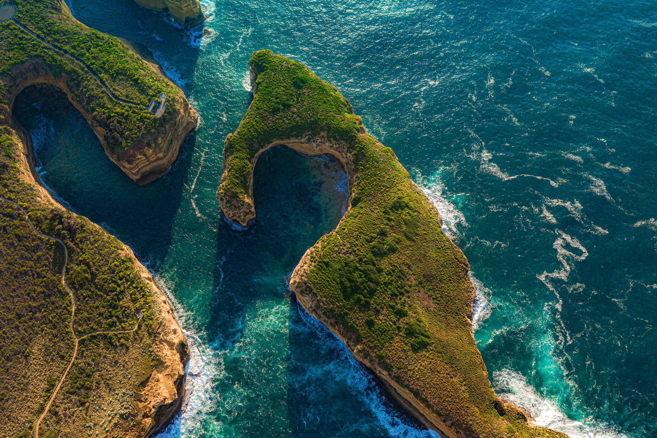 Aerial view of a coastal area with sea arches and cliffs covered in greenery, with turquoise water surrounding the formations.