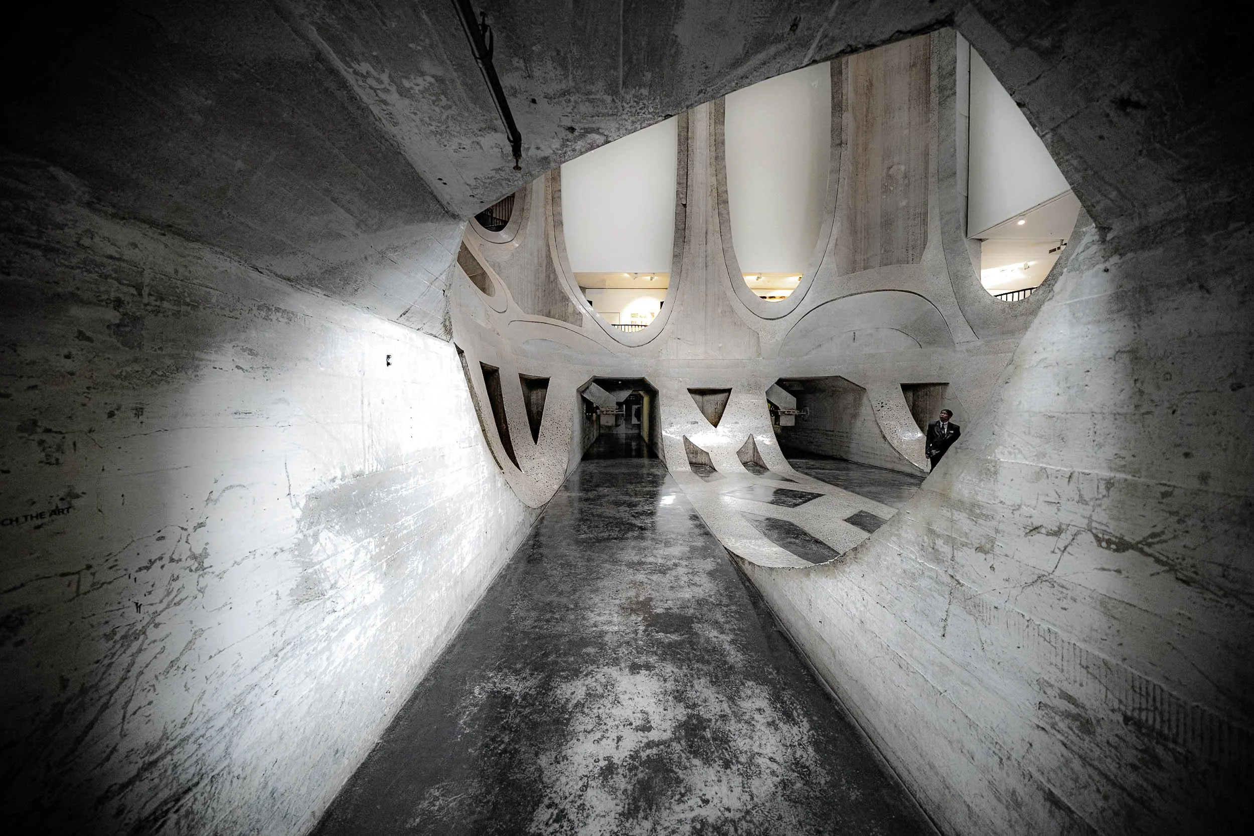 A modern interior with concrete walls and unique architectural design featuring large, curved openings and a person standing in the corner.