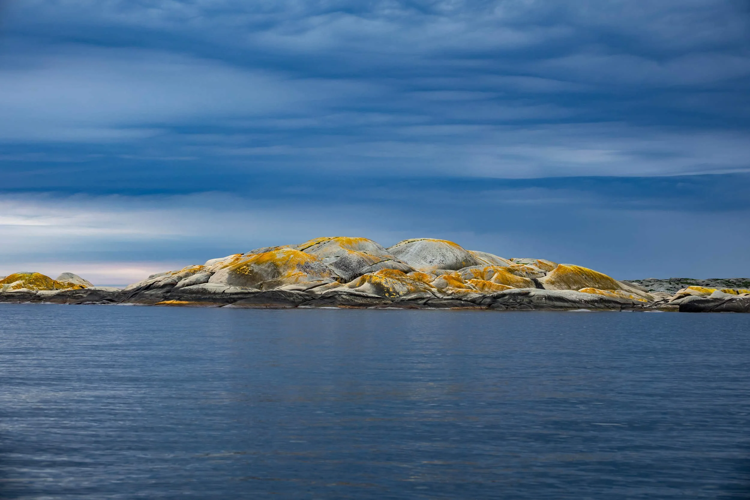 Rocky island with moss and lichen in a body of water under a cloudy sky.