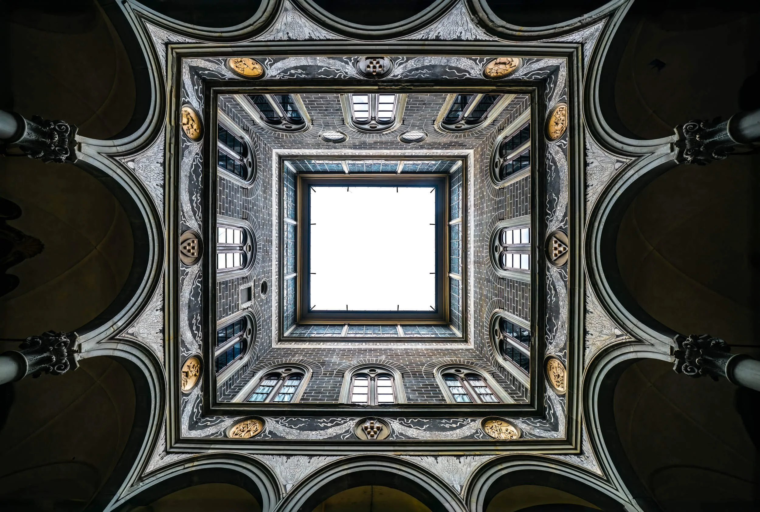 Looking up at a multi-story building's interior courtyard with an open skylight at the top, surrounded by ornate arches and detailed decoration.