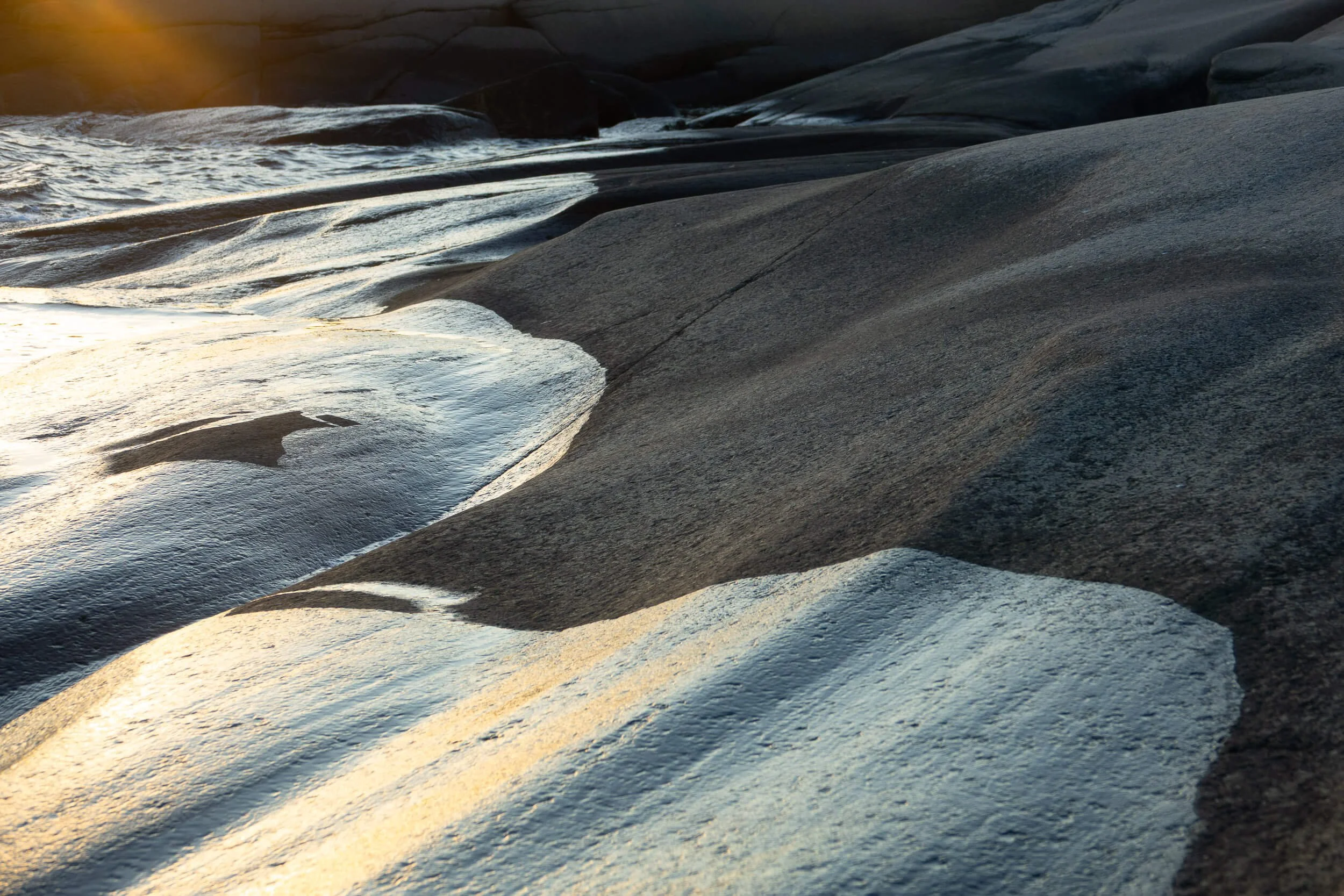 Close-up of smooth, layered rocks along the shoreline with gentle water flow, illuminated by warm sunlight.