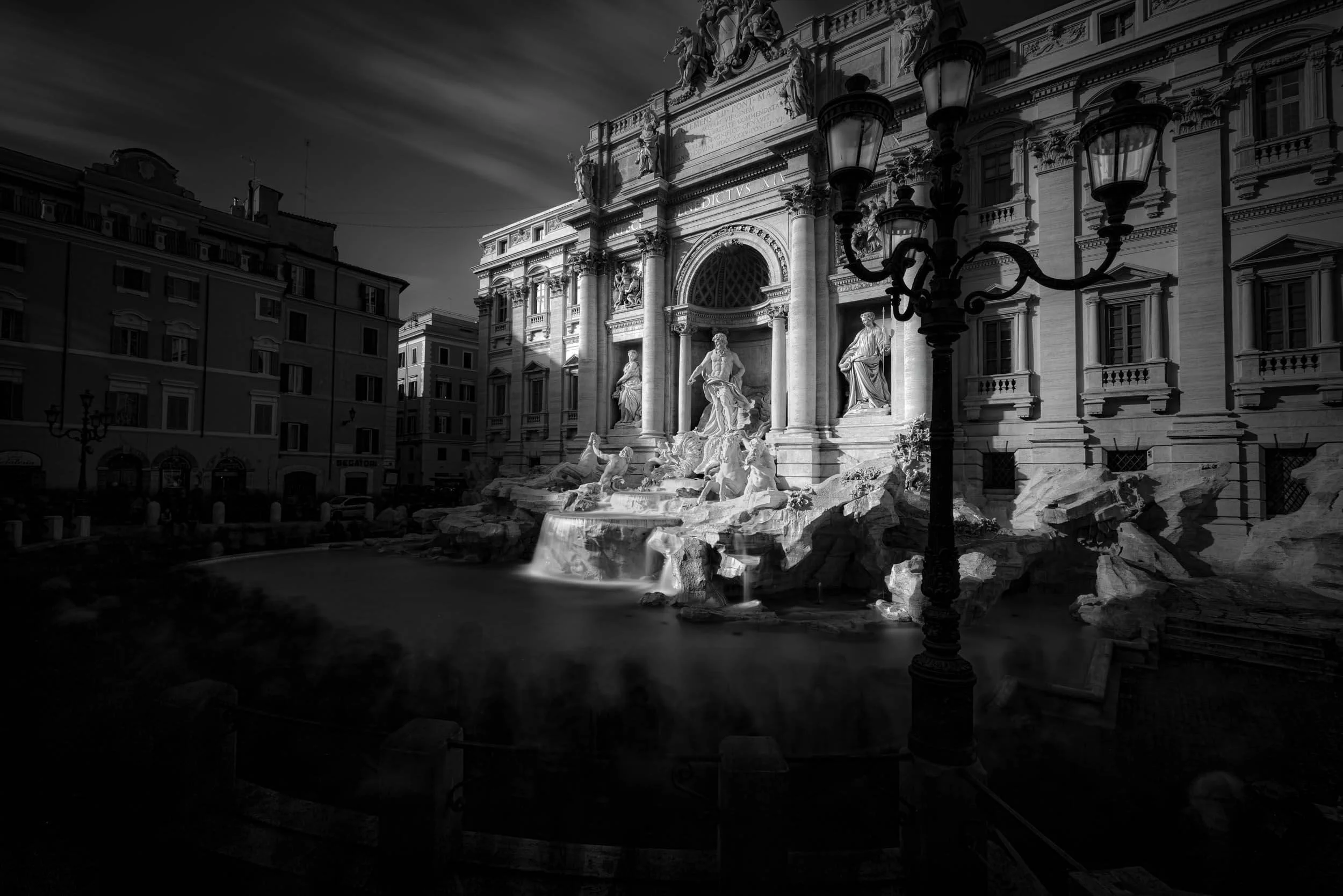Black and white photo of a historic European-style building with ornate architectural details, statues, and a fountain in front. A street lamp is visible near the fountain.
