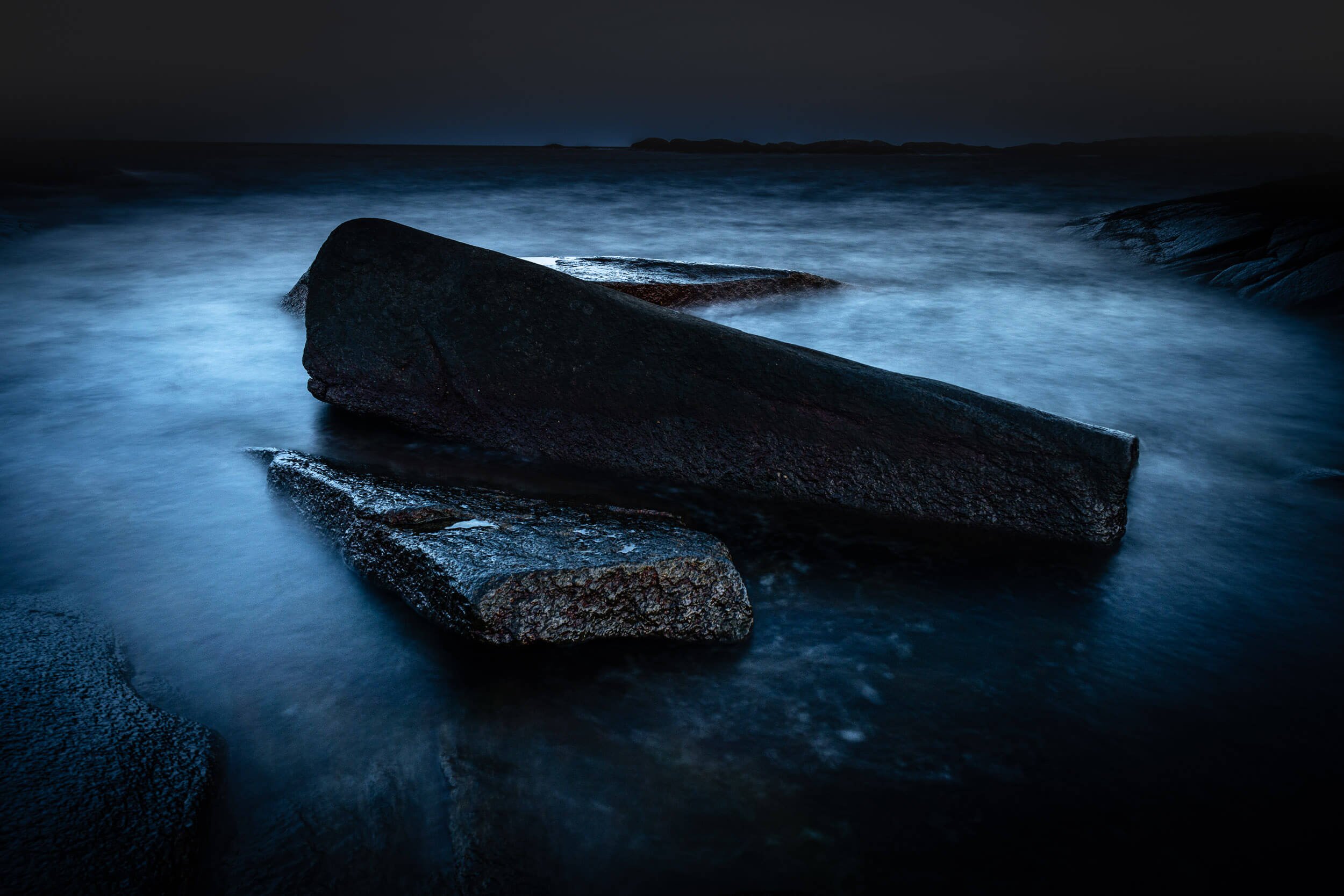 Dark night scene of ocean waves crashing against large rocks along the shoreline with a distant landmass on the horizon.