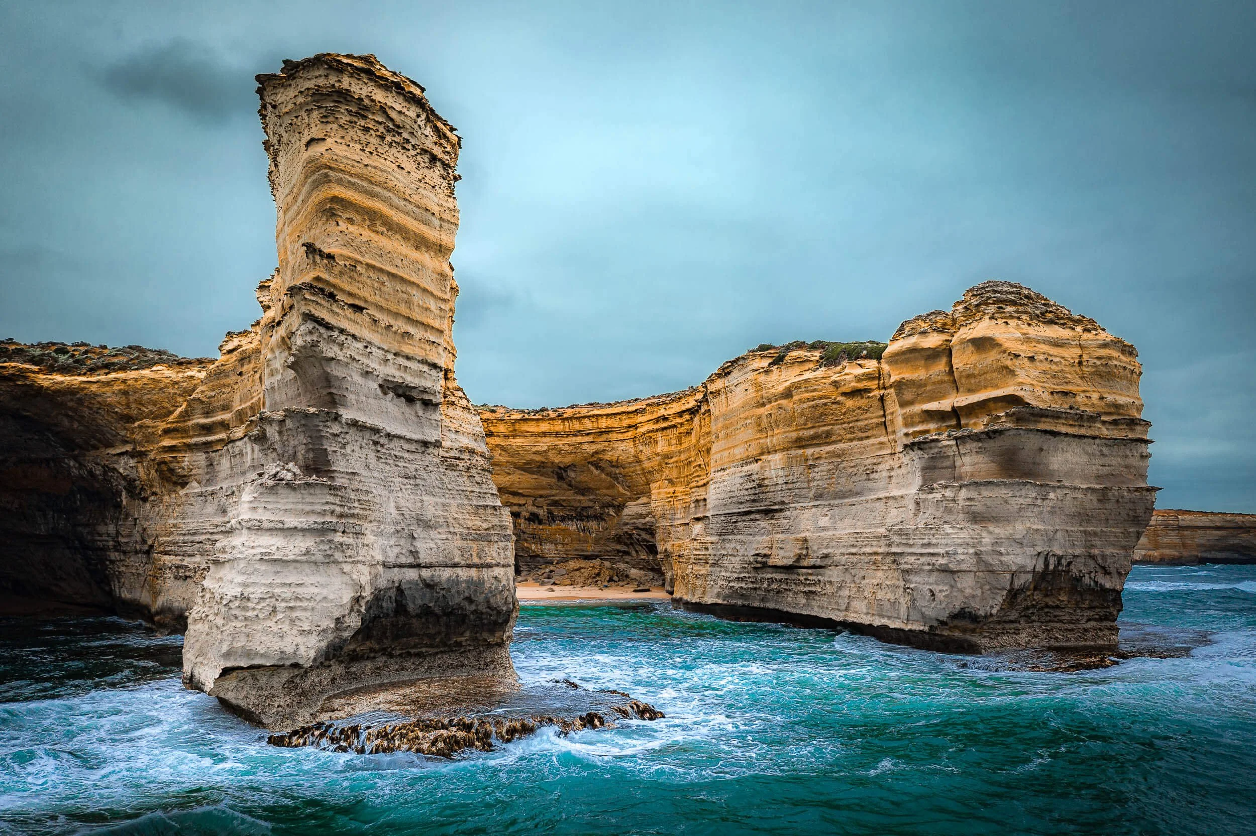 Cliffs and rock formations along a coastline with waves crashing at the base, under a cloudy sky.