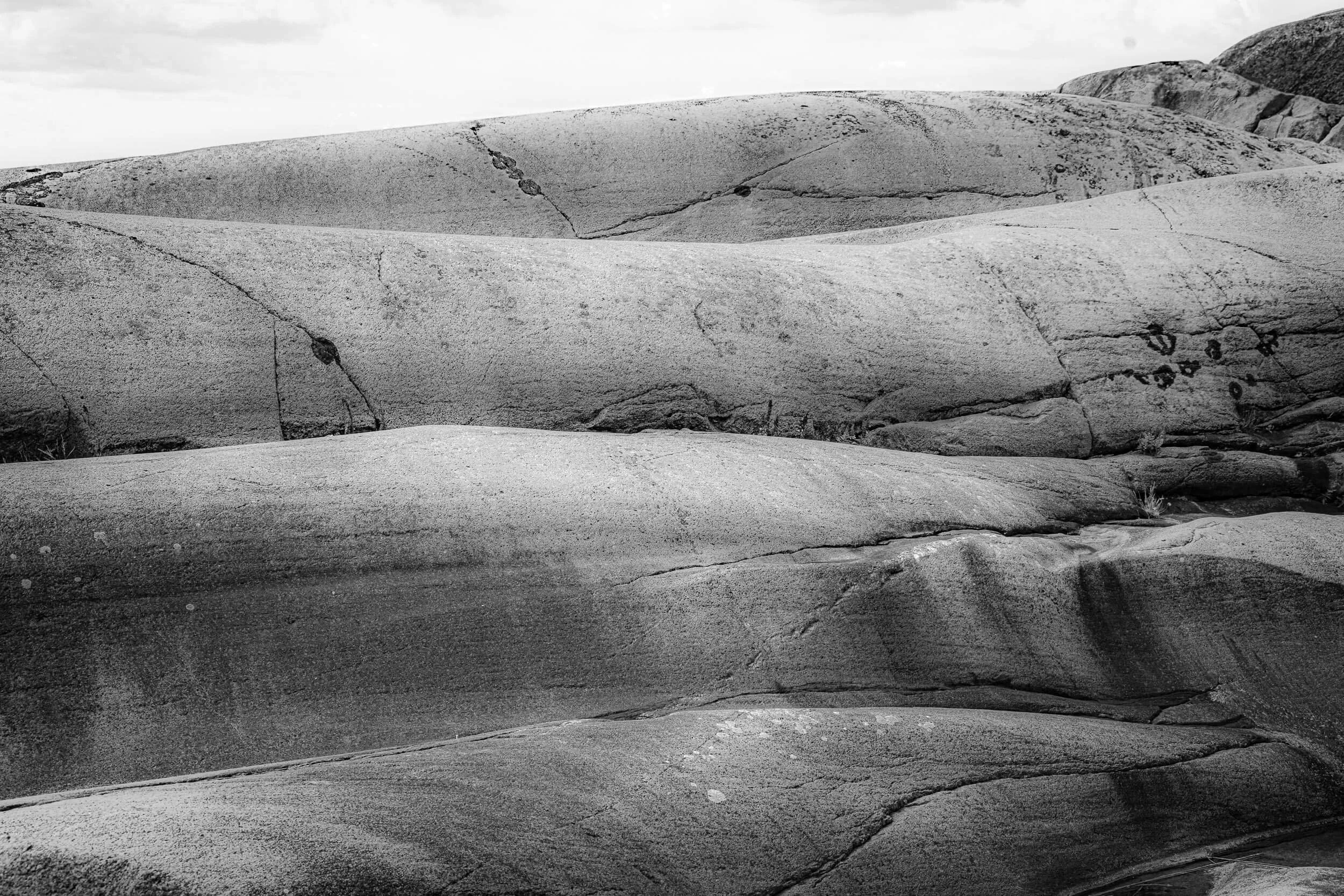 Black and white photograph of large smooth rocks with visible cracks and sparse vegetation, under a cloudy sky.
