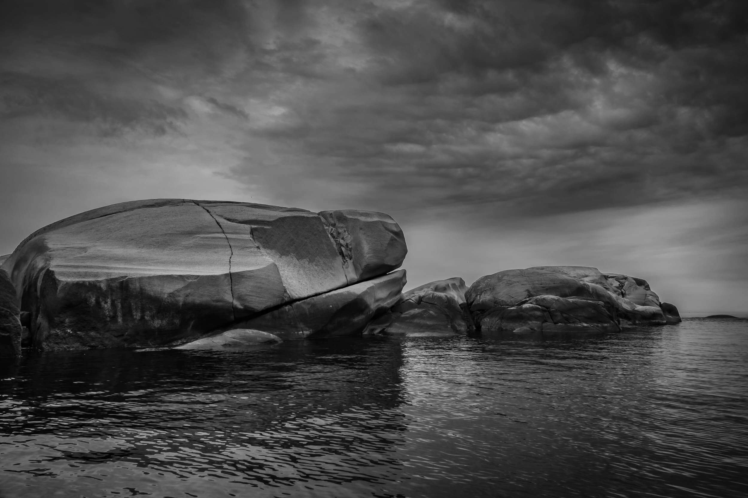 Black and white photo of large smooth rocks along a body of water with clouds overhead.