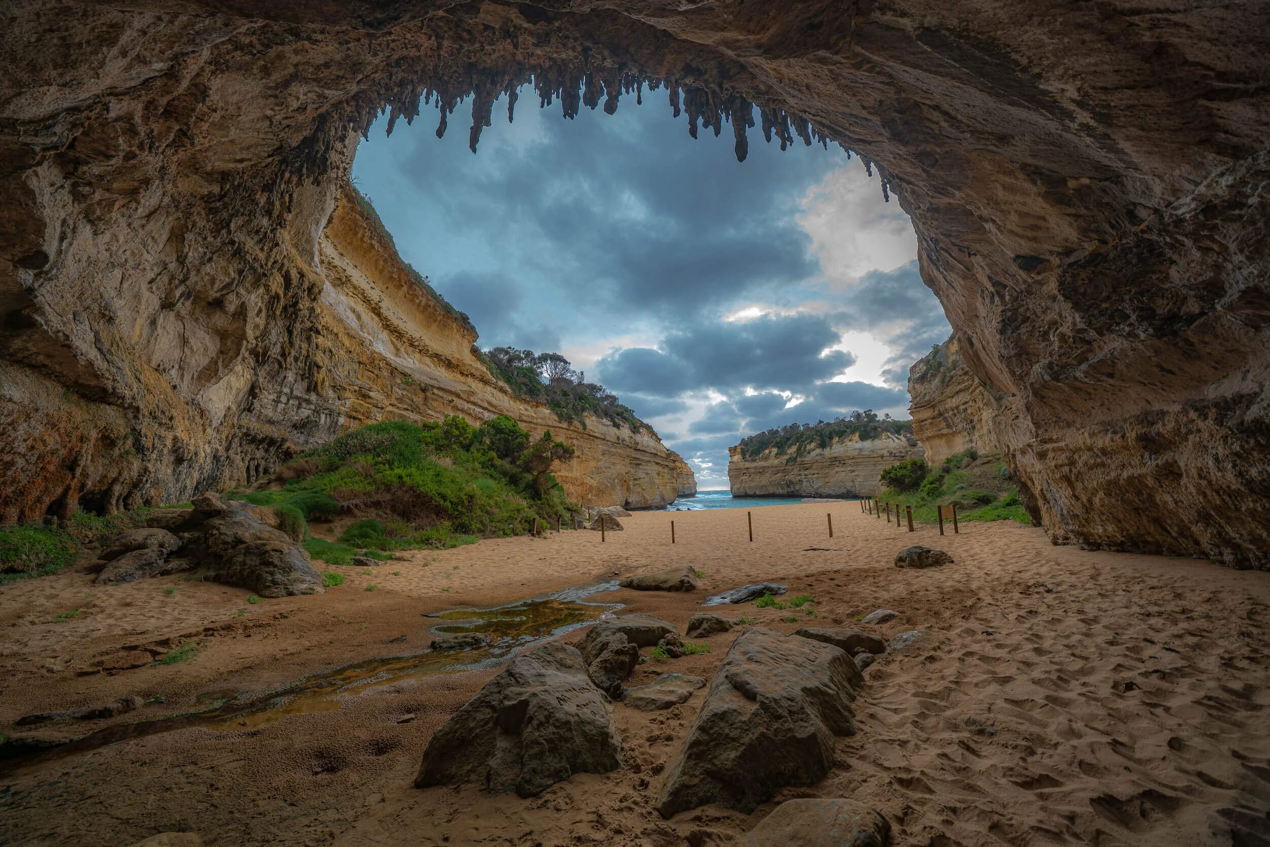 View of a beach seen from inside a large cave with rocky walls and a sandy floor, with some greenery and ocean visible in the distance under a cloudy sky.