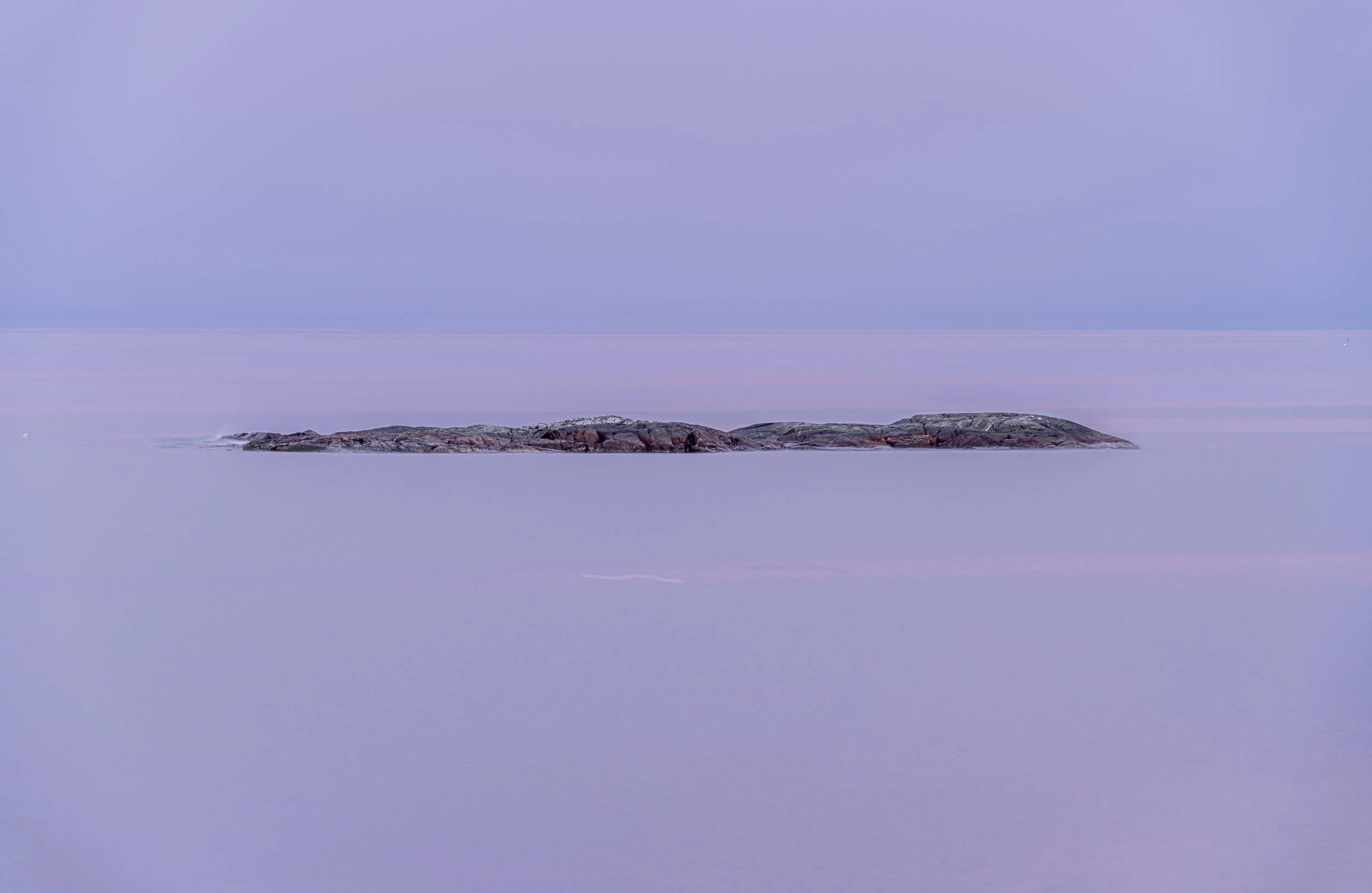 A calm sea with a small rocky island in the foreground, under a pastel-colored sky with soft clouds.
