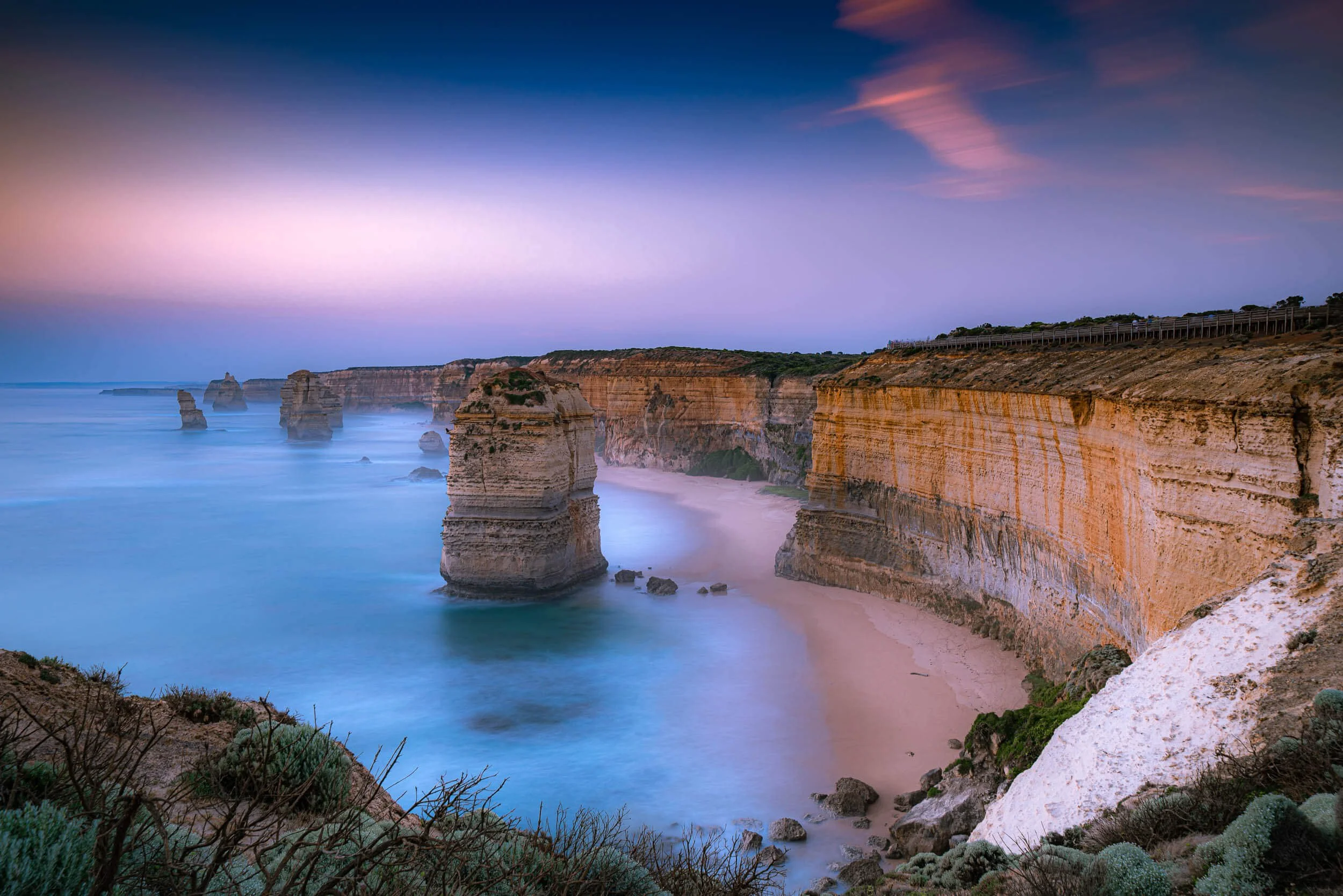 Cliffs along a rocky coastline with tall sea stacks, sandy beach, and a colorful sky at dusk.