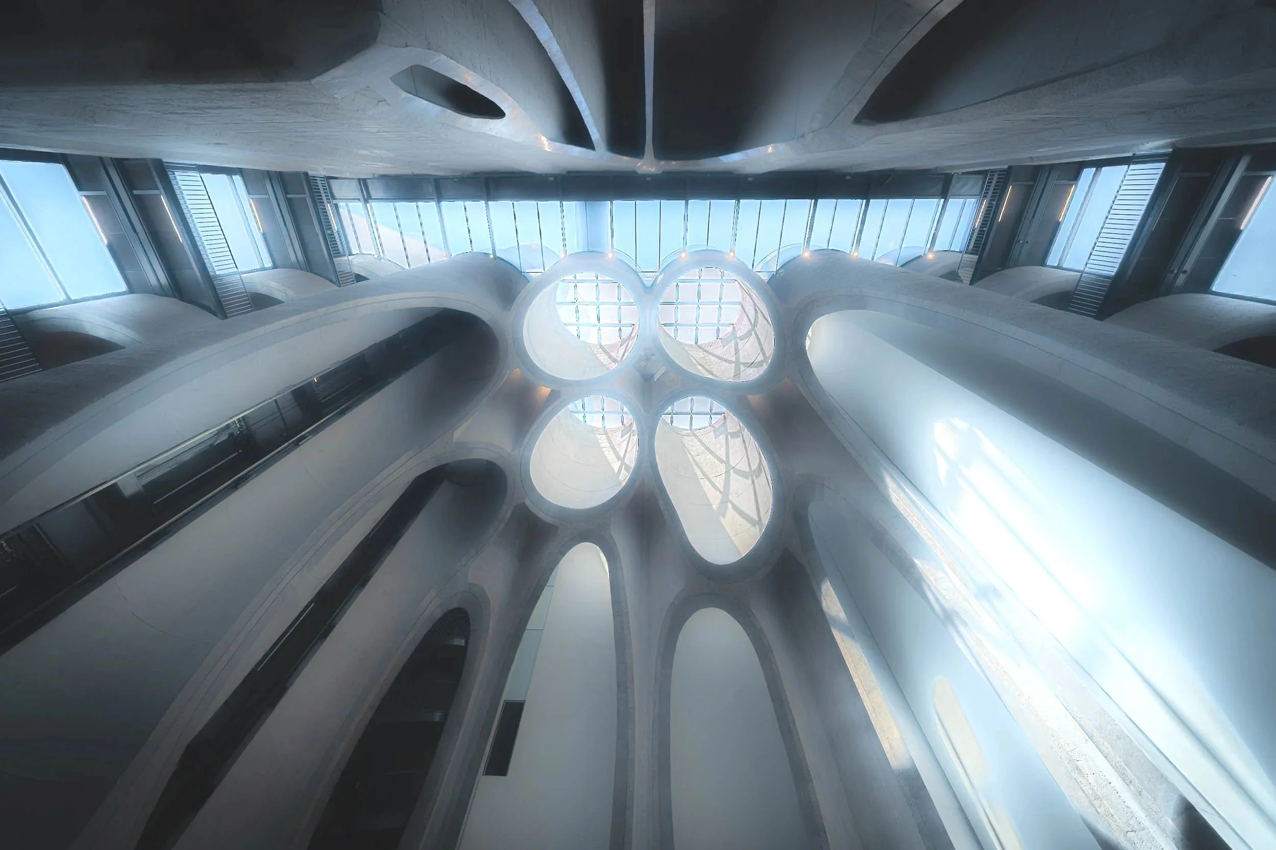 View of a modern indoor space with tall, curved architectural features and large glass windows allowing natural light, looking upward from the ground floor.