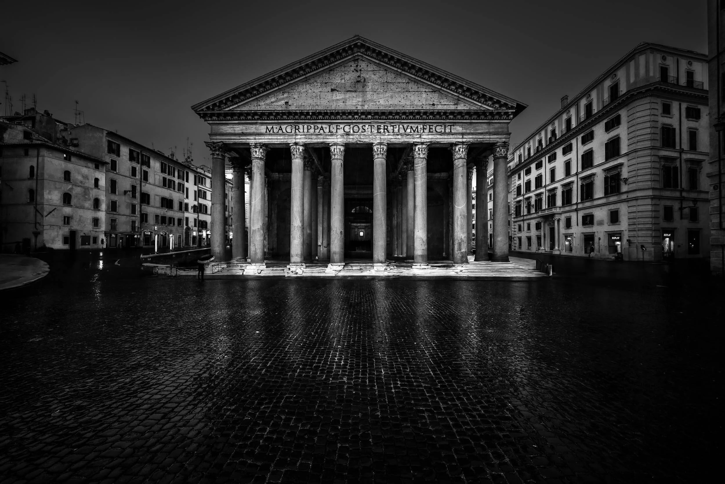 Black and white photo of the ancient Pantheon building in Rome with classical columns and Latin inscription, surrounded by modern buildings, wet cobblestone ground, likely after rain.