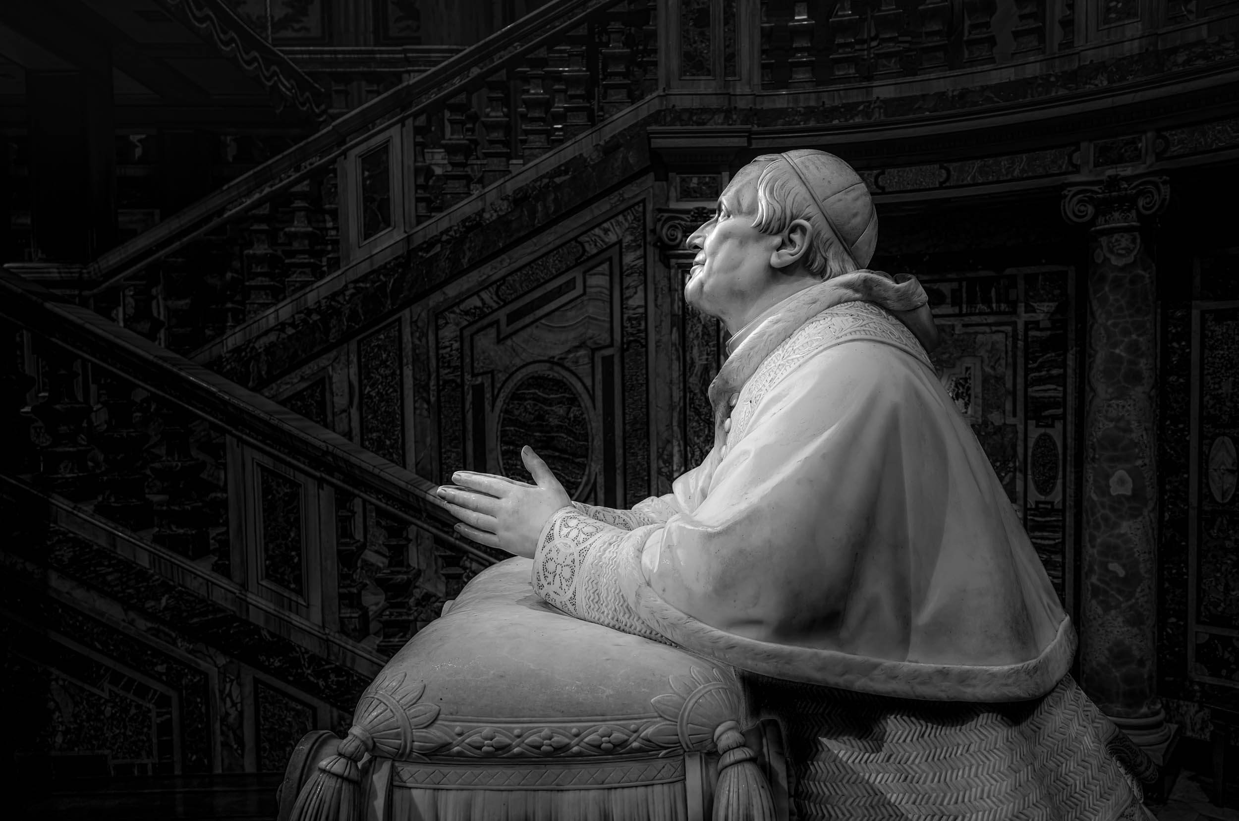 Black and white photograph of a detailed sculpture of a sitting man, who is smiling and wearing traditional clothing, in front of an ornate staircase.