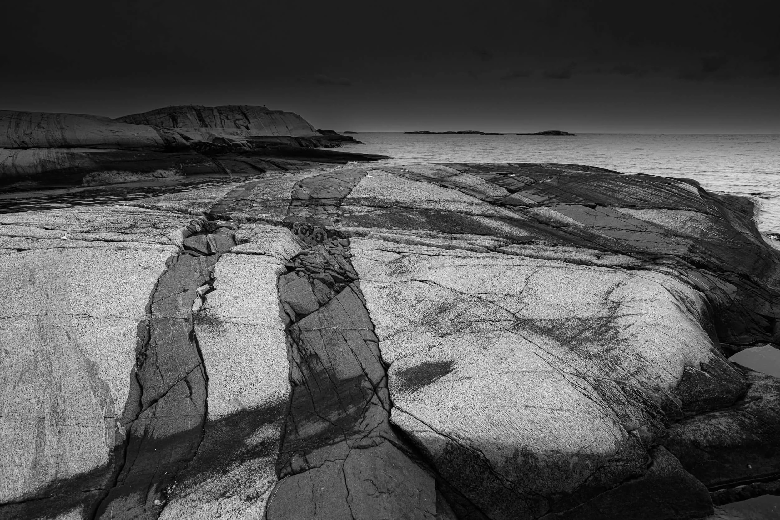 A black and white photograph of a rocky coastal landscape with large granite formations, a calm sea, and a cloudy sky in the background.