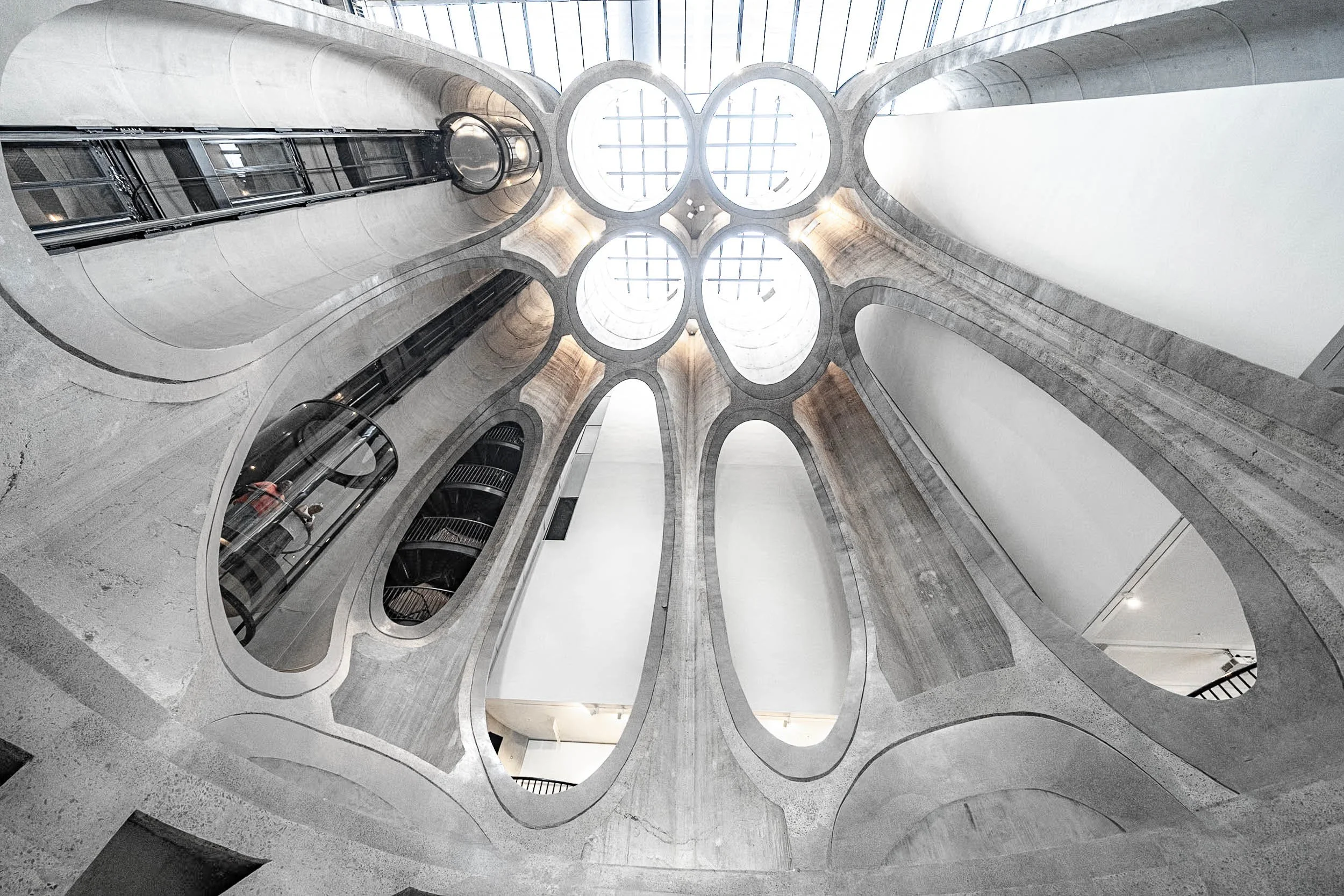 Interior view of a modern building with multiple spiral concrete staircases and a glass ceiling allowing natural light inside.