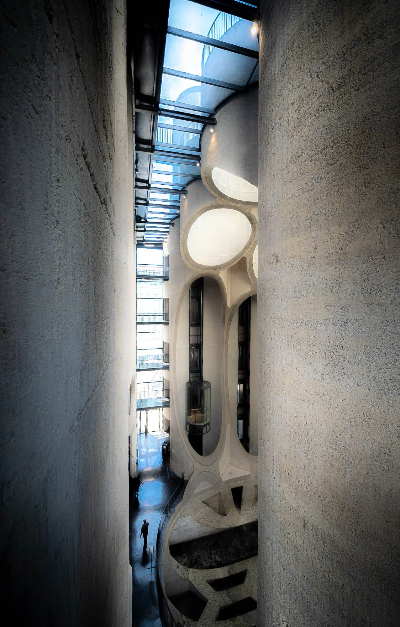 Interior view of a modern building with white curved walls, large oval openings, and a spiral staircase. A person is walking at the bottom of the space, with natural light coming through windows and glass ceiling panels.