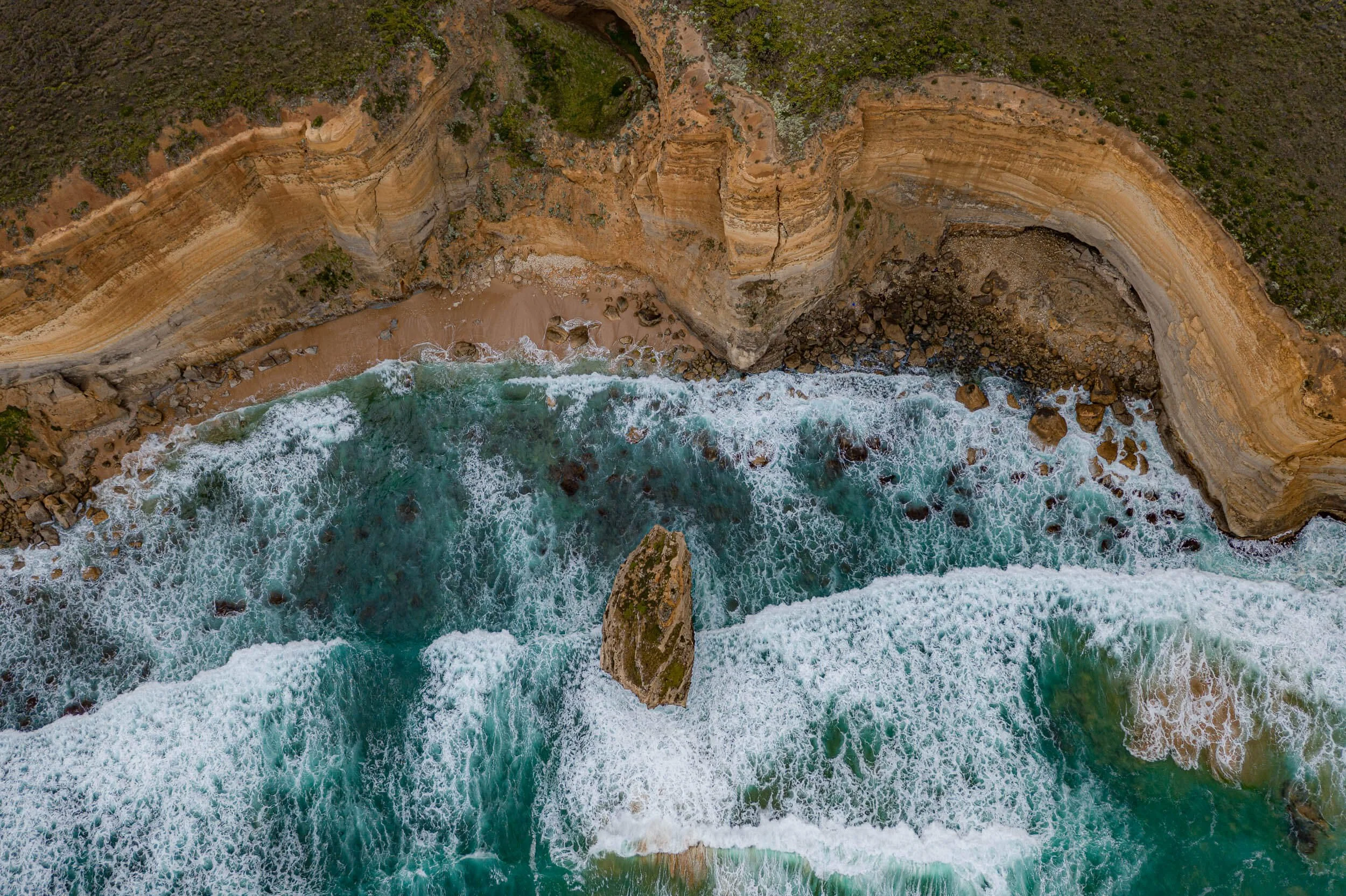 Aerial view of a coastal scene showing eroded cliffs, a sandy beach, and turquoise ocean waves crashing against rocks.