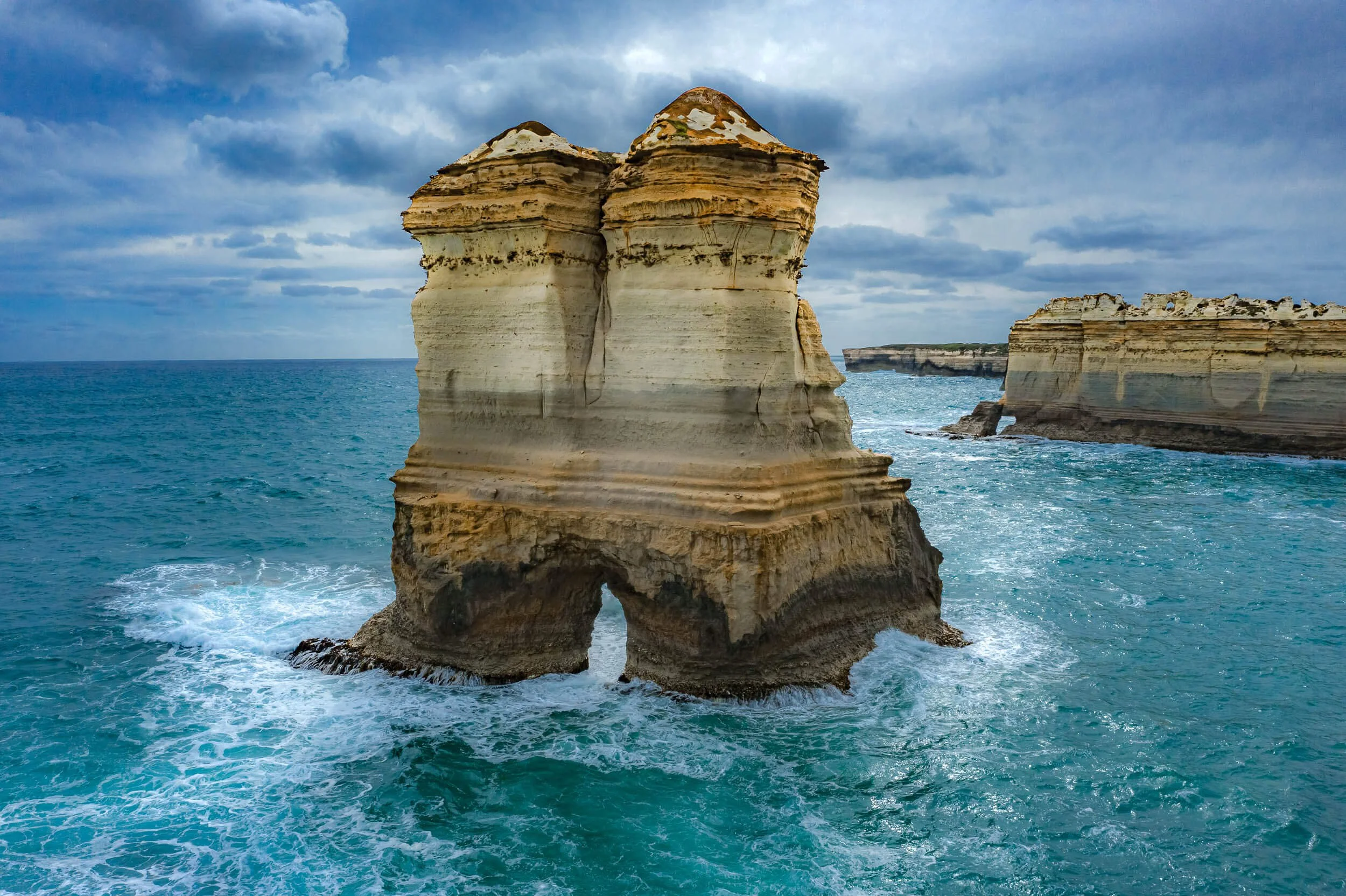 Large limestone rock formations jut out of the ocean, with the main formation having a natural arch at its base. The sea is blue-green with white waves at the base of the rocks. The sky is mostly cloudy with patches of blue.