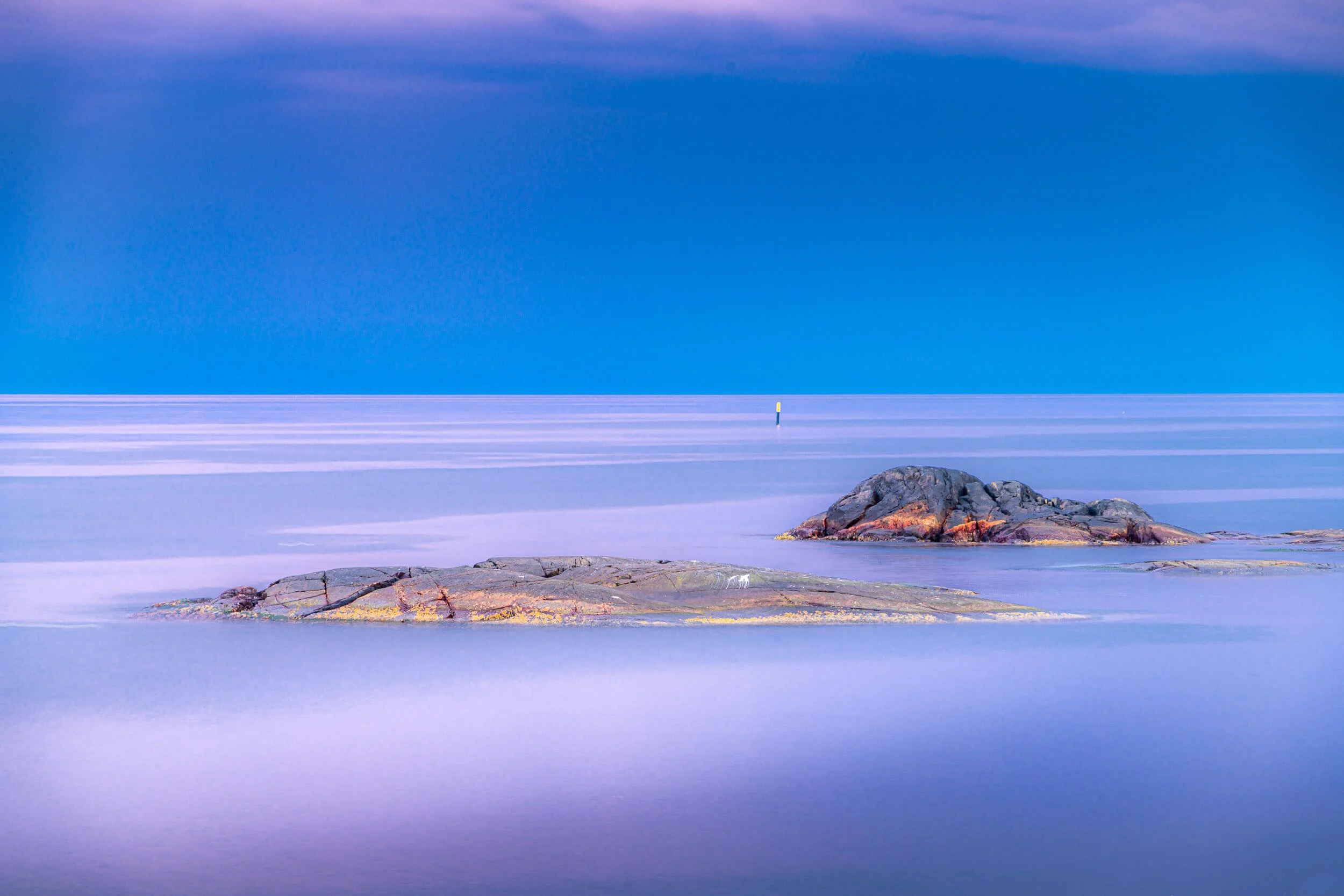 Calm ocean with a small rocky island, a buoy in the water, and a hazy purple and blue sky.