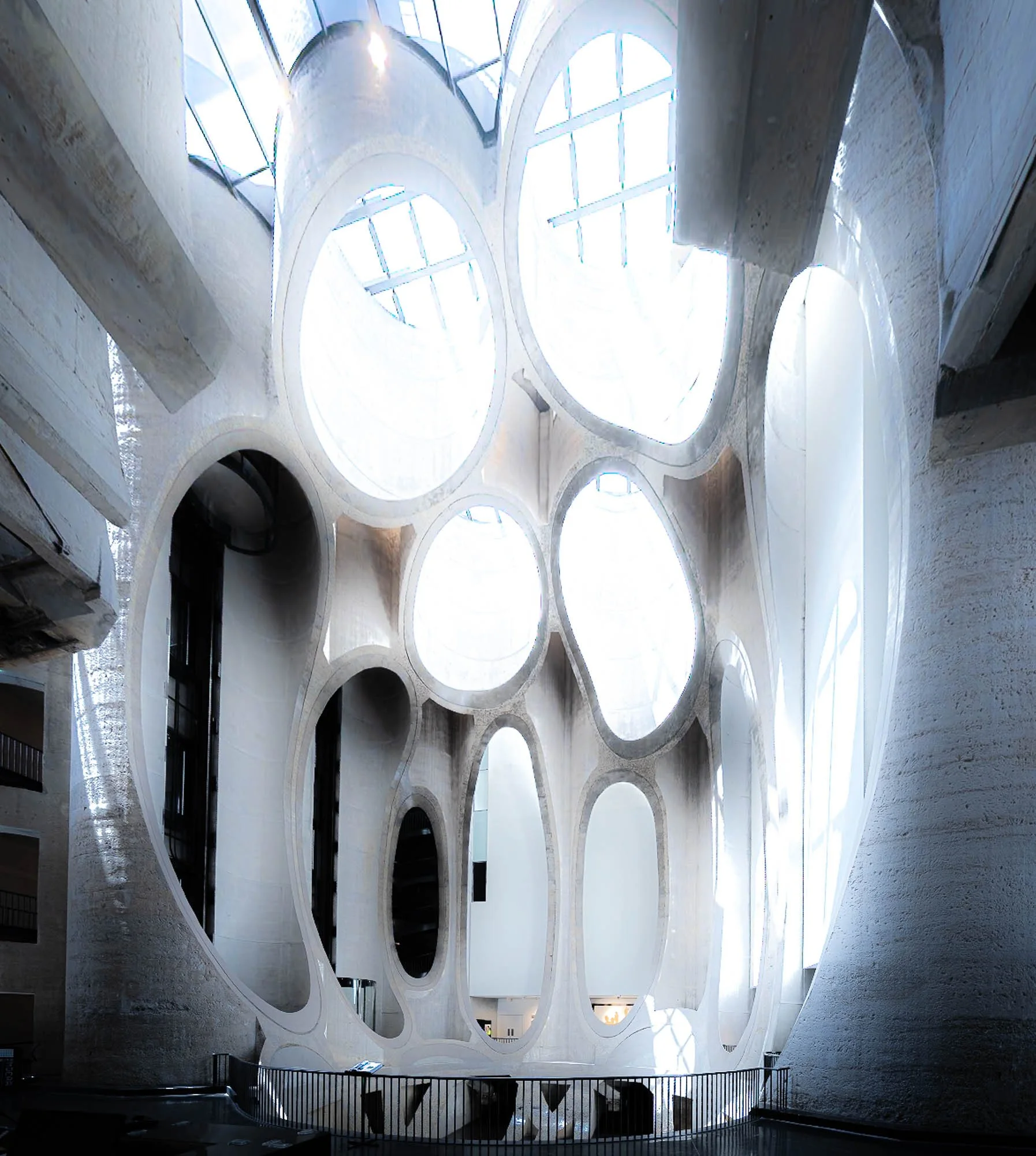 Interior view of a building with large white, organic-shaped openings in the ceiling, allowing natural light to enter, with a viewing balcony and stairs.