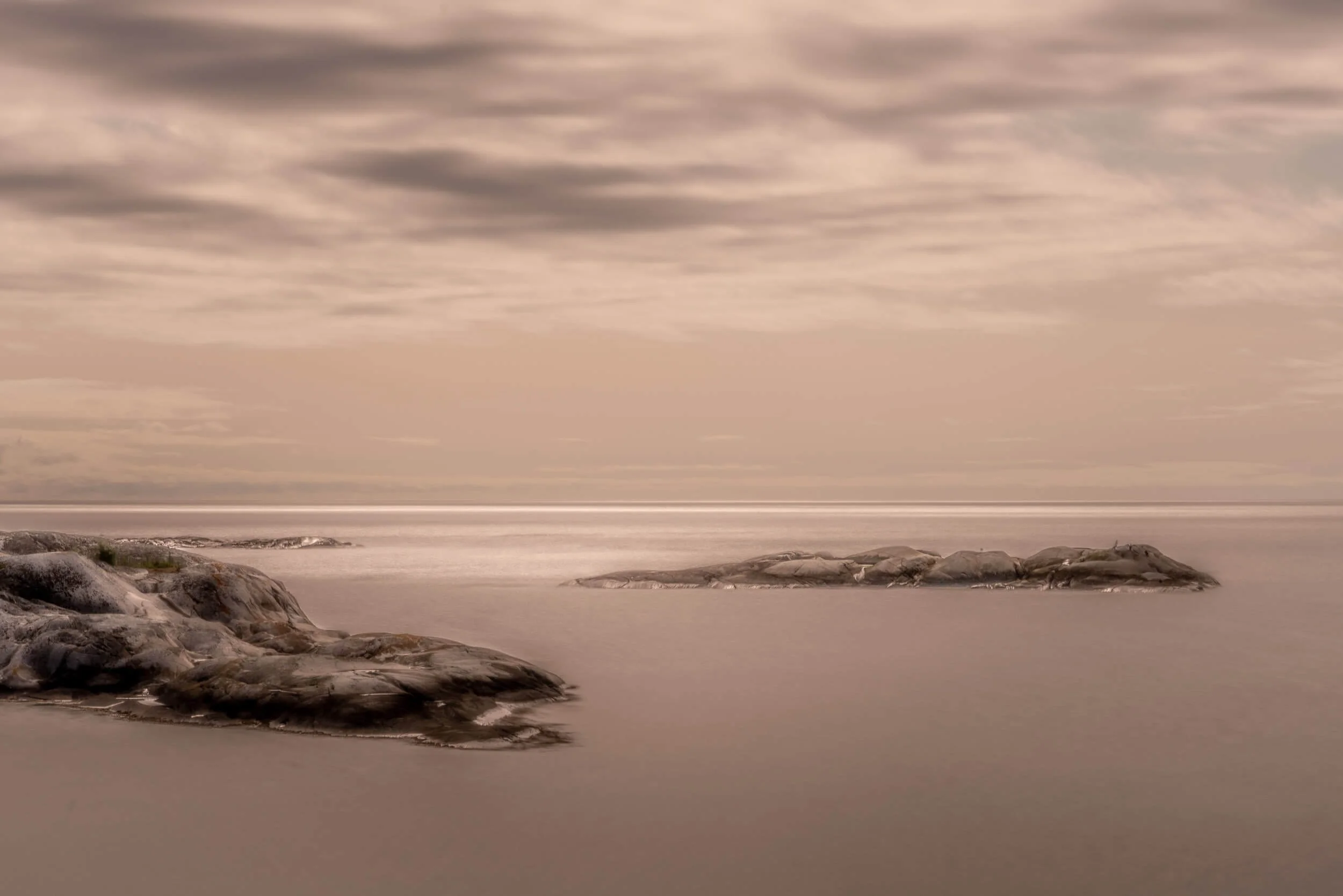 A tranquil seascape scene featuring smooth rocks extending into calm water under a sky filled with soft, layered clouds during sunset or sunrise.