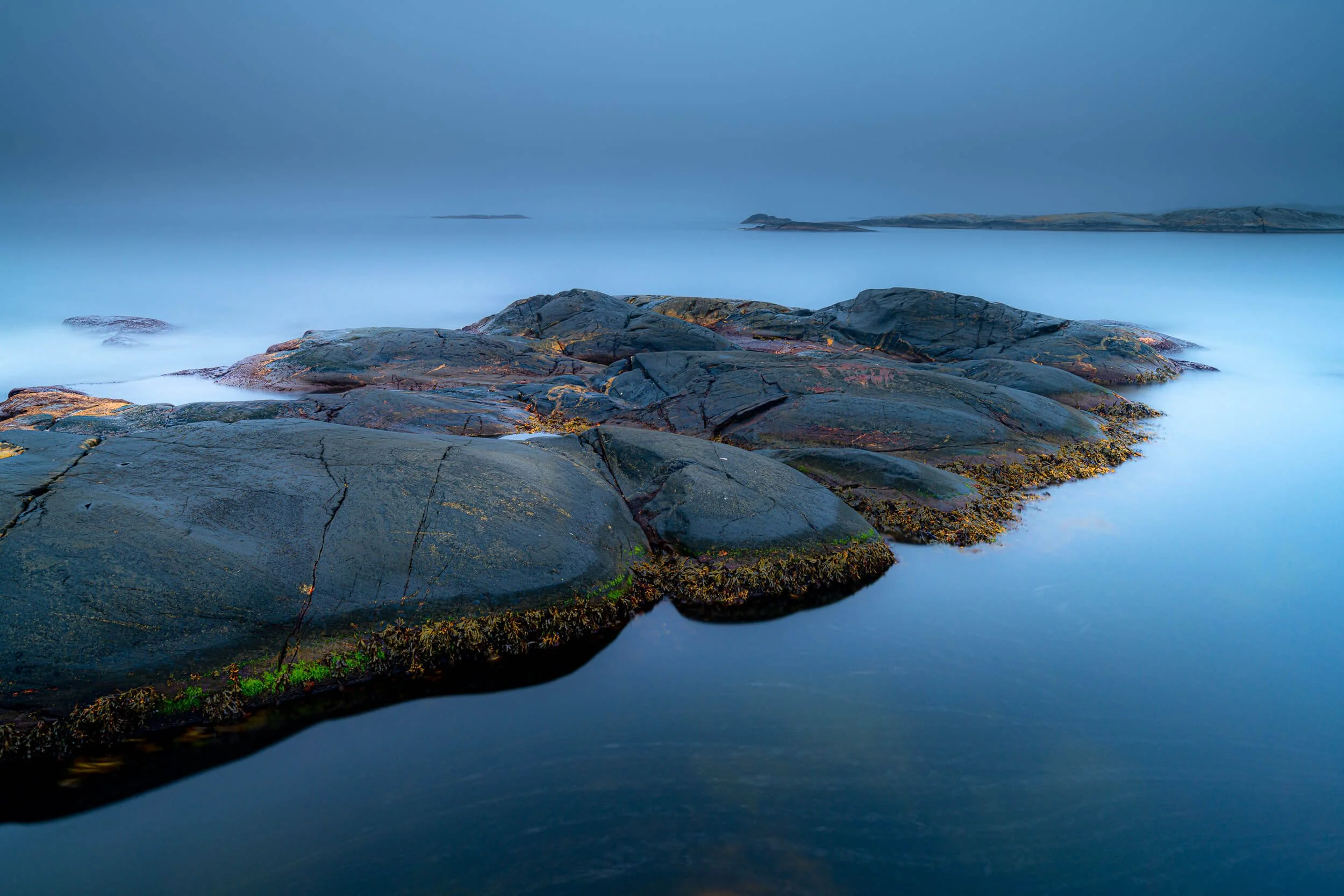Smooth ocean water surrounding large, dark rocks along the shoreline with distant land and a cloudy, overcast sky in the background.