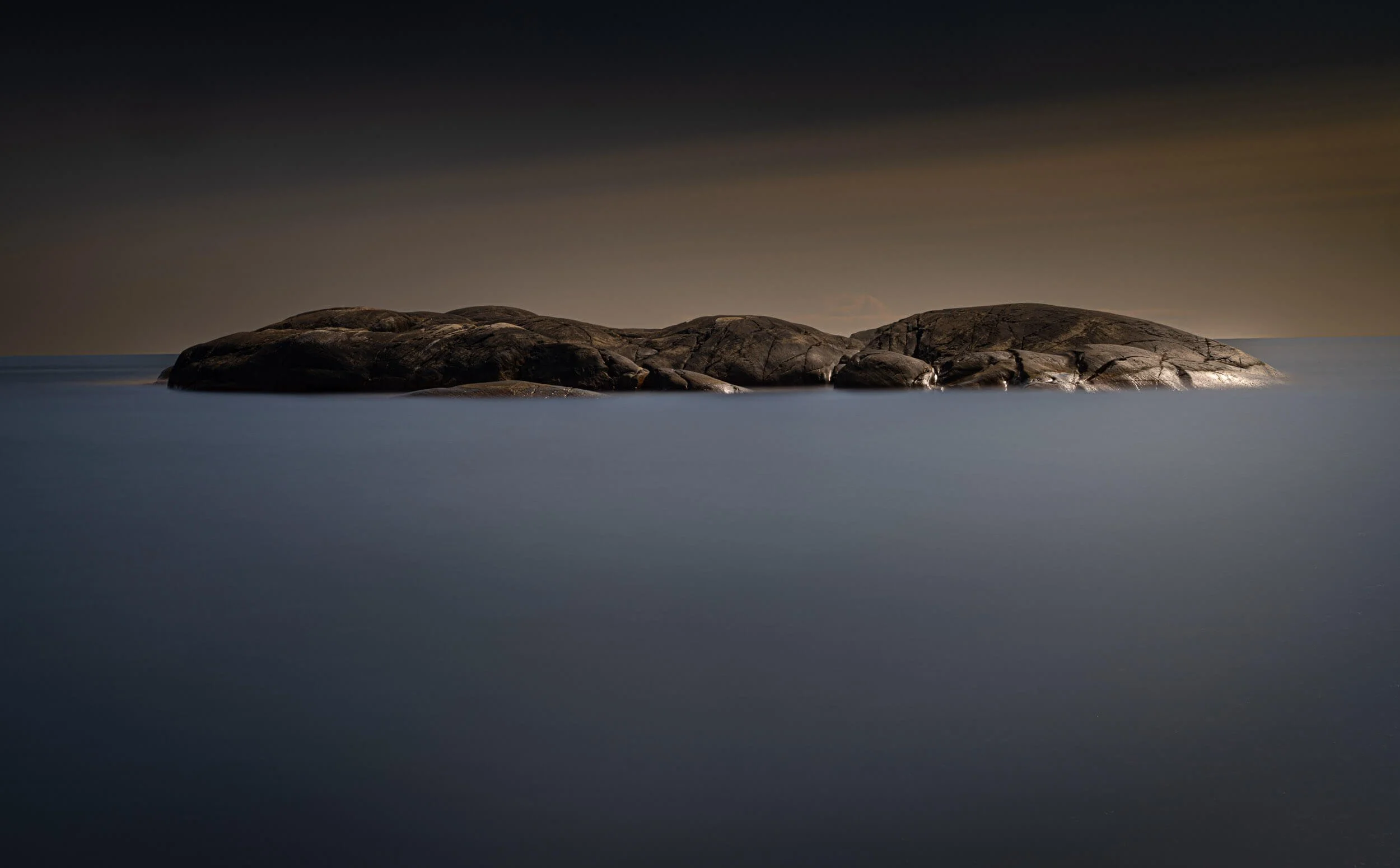 A rocky island surrounded by calm water during dusk or dawn, with a dark sky overhead.