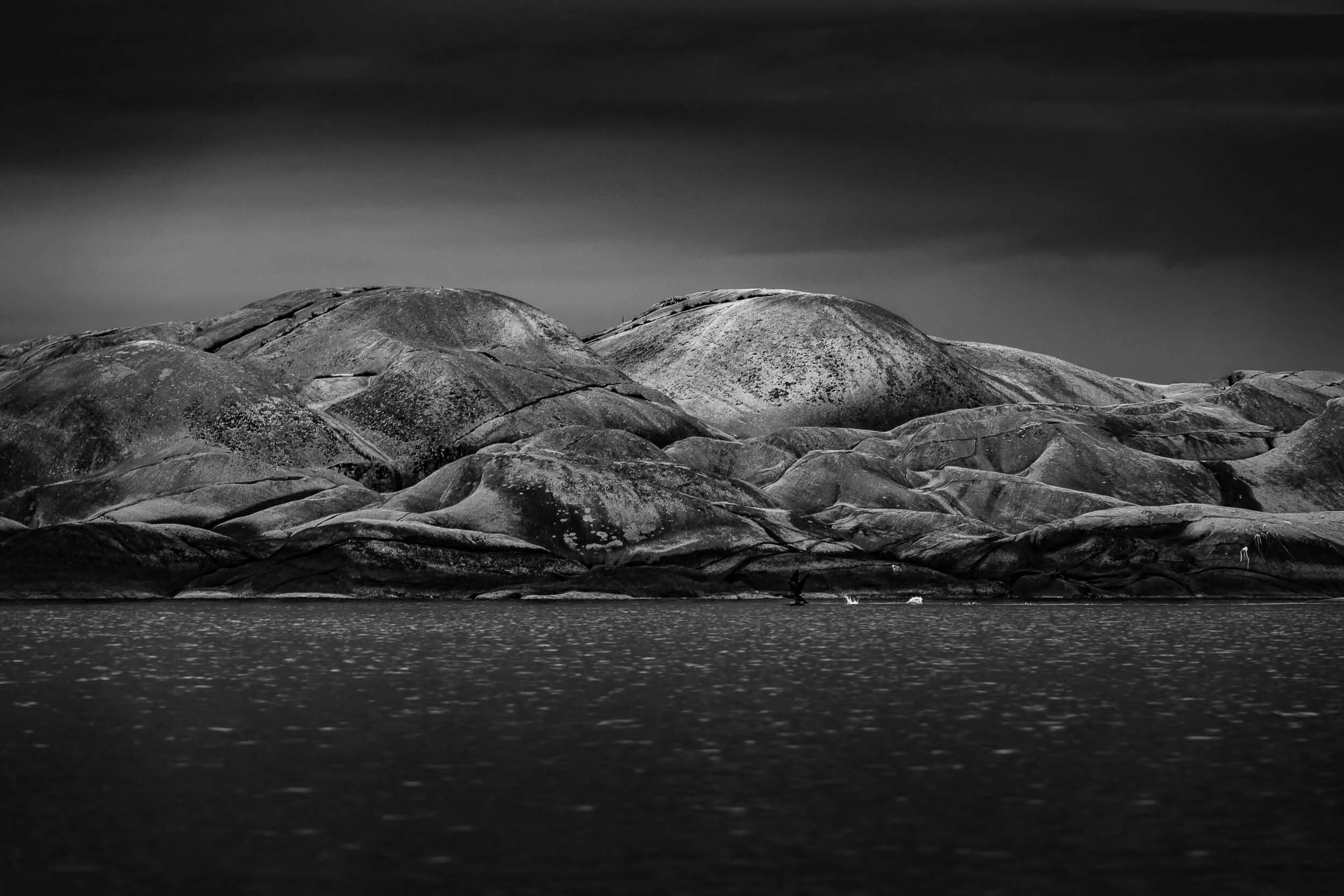 Black and white photo of large, smooth rocks or boulders by a body of water, with a person in a boat near the shore and dark clouds in the sky.