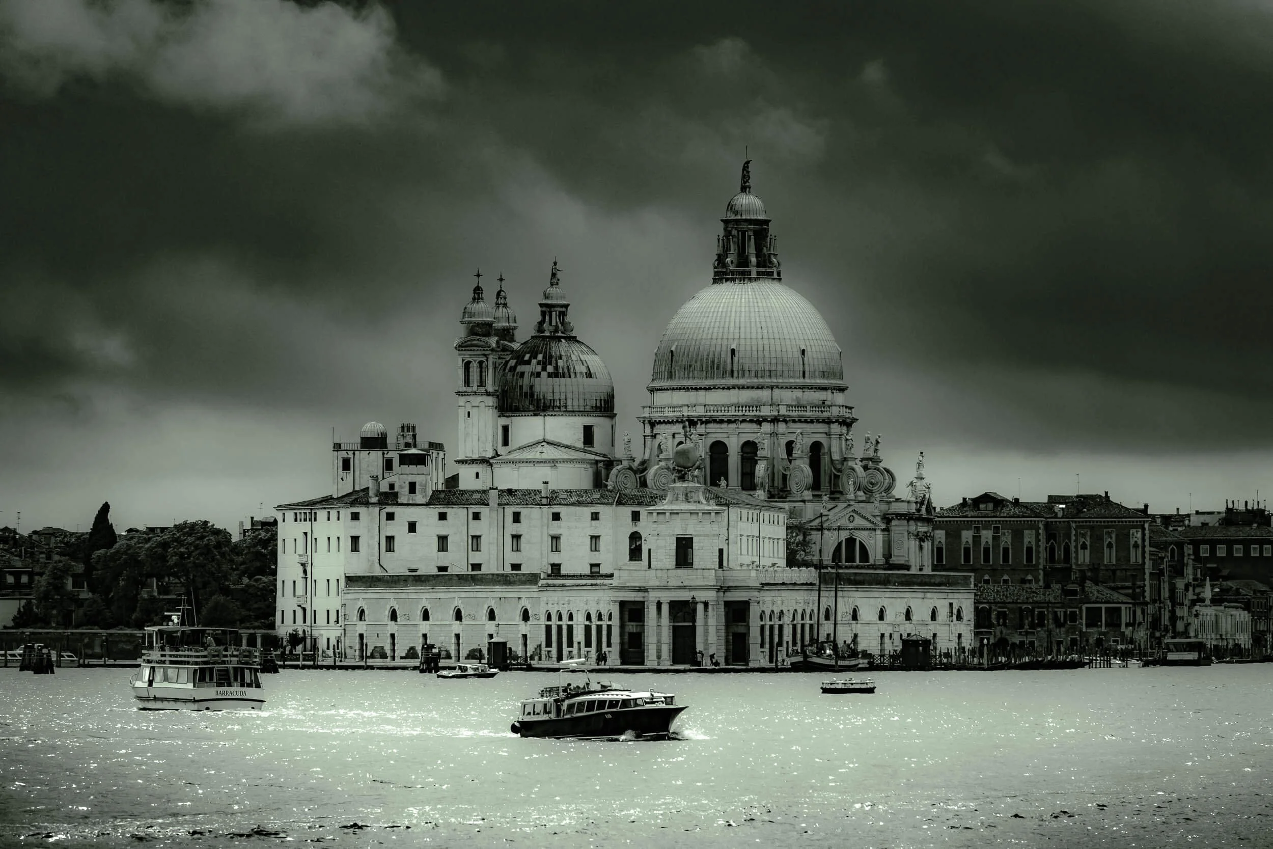 View of historical buildings with domes and a large central dome topped with a statue, near a body of water with boats, under a dark stormy sky.