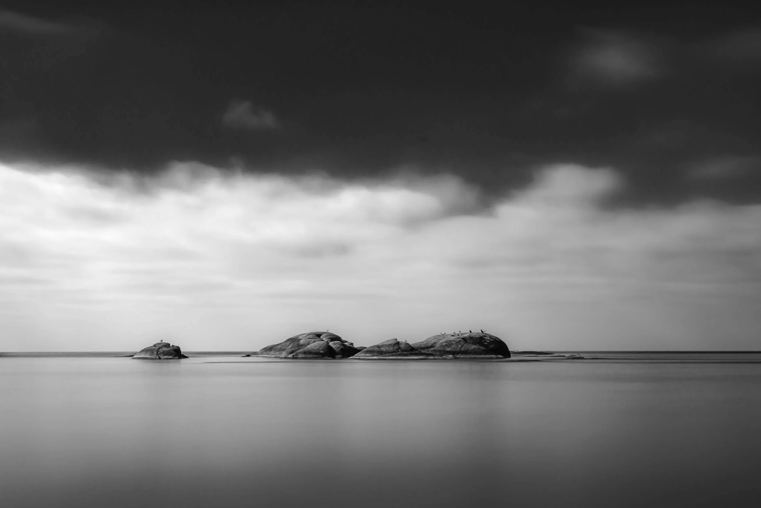A black and white photograph of a rocky shoreline with boulders in calm water and a cloudy sky above.