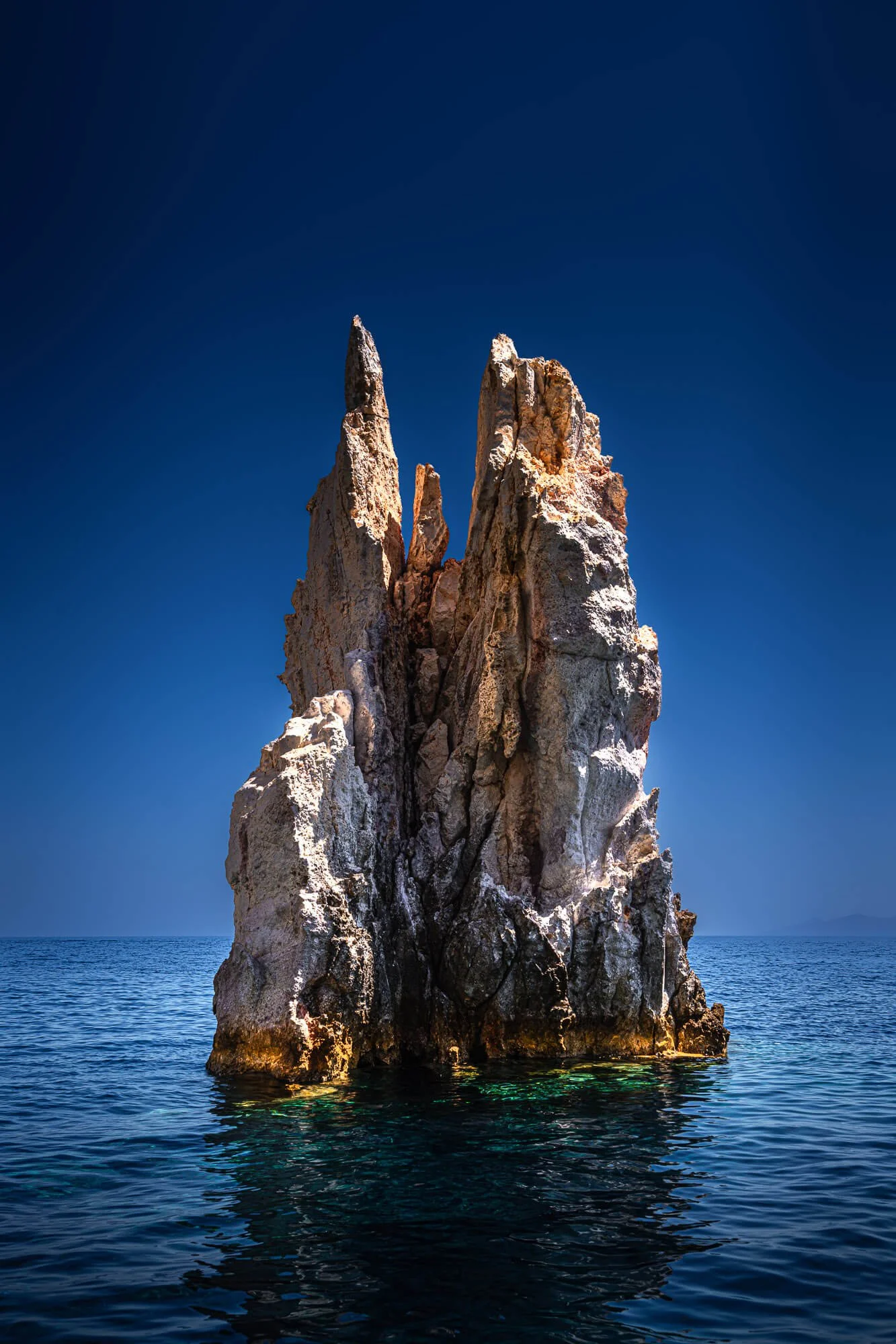 A tall rocky sea stack rises from the ocean with a clear blue sky above and calm water below.