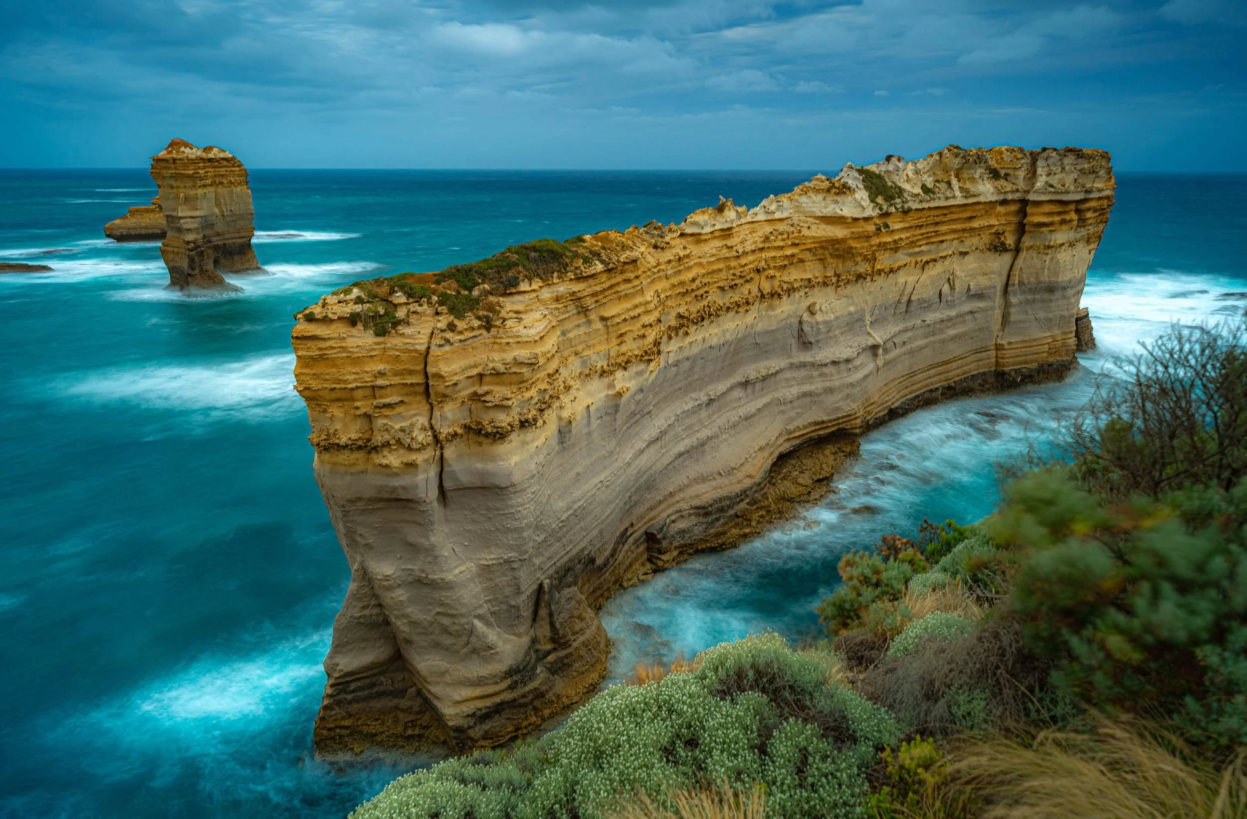 Ocean cliffs with layered rock formations and a sea stack, under a cloudy sky.