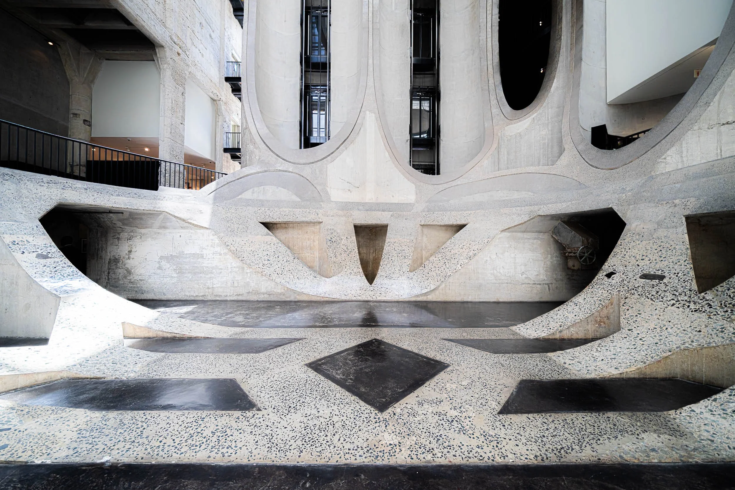 Interior of a modern building with curved concrete walls, large openings, and an elevator shaft. The design features smooth, flowing lines and a speckled terrazzo floor in black, white, and gray.